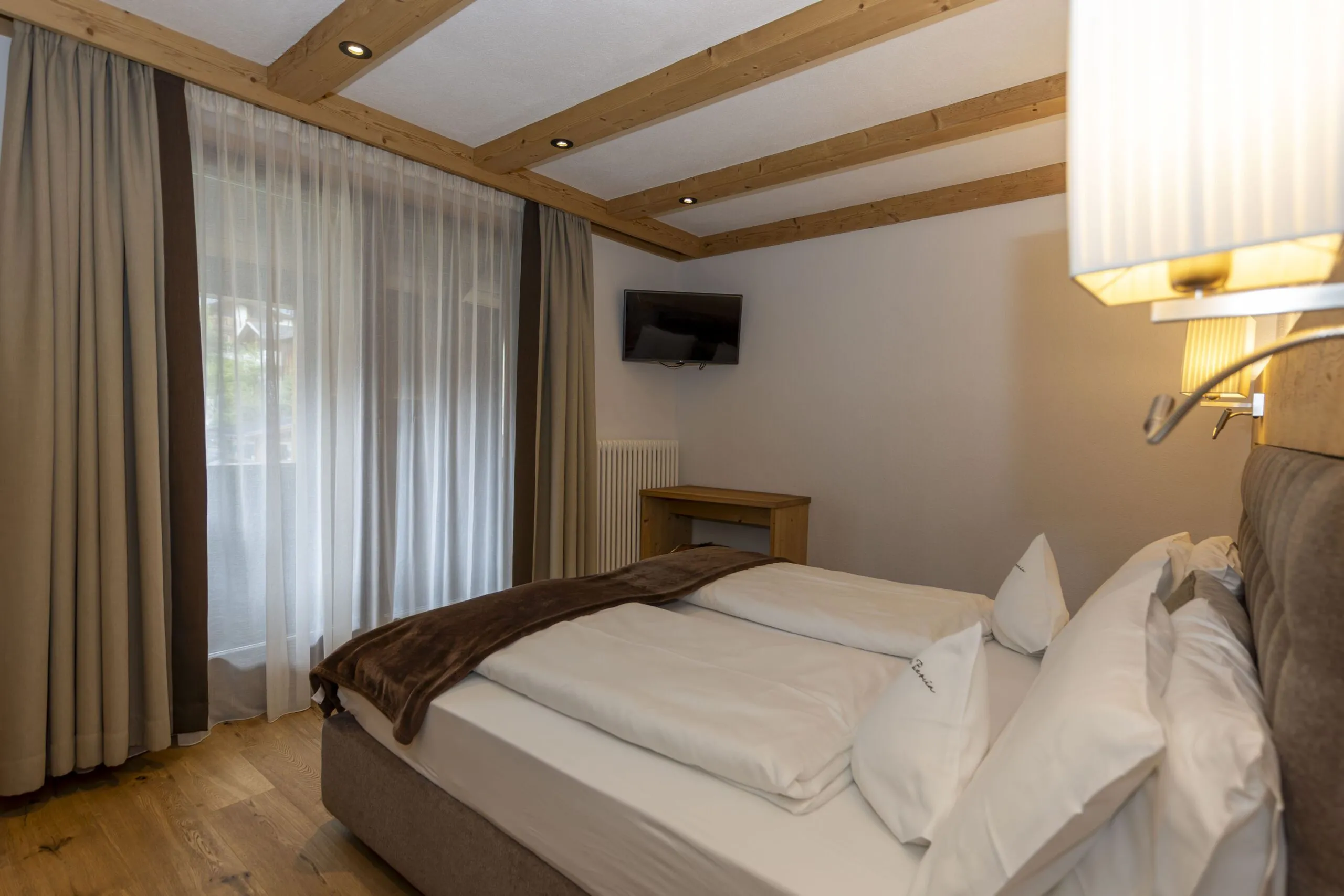 Hotel bedroom with a double bed, wooden beams ceiling, sheer and beige curtains, wall-mounted TV, and small desk.