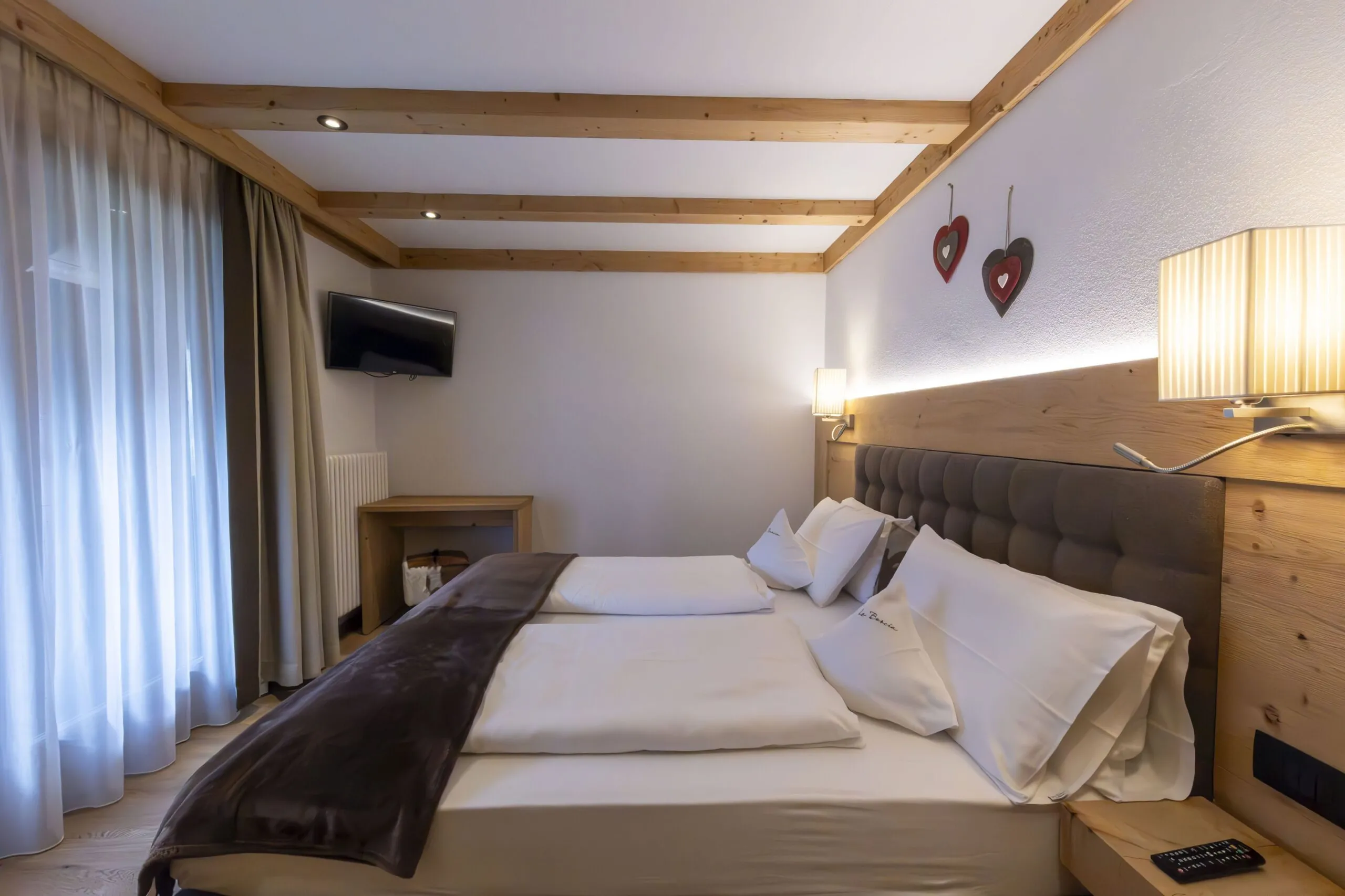 Cozy hotel bedroom with a large bed, wooden beams, wall-mounted lamps, and a hanging TV.