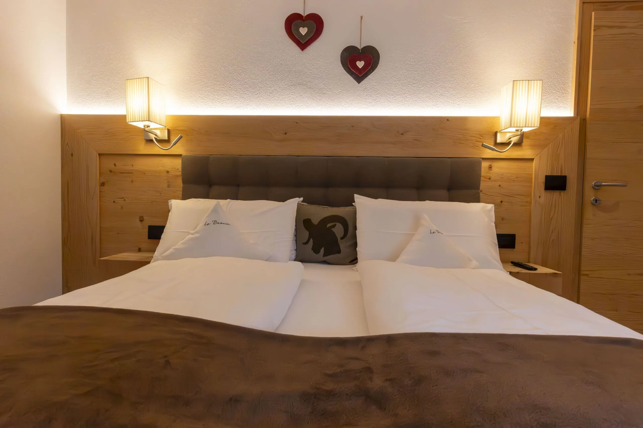 Cozy hotel bed with white linens, brown blanket, tufted headboard, wooden accent wall, two wall lamps, heart wall decor above, and side tables.