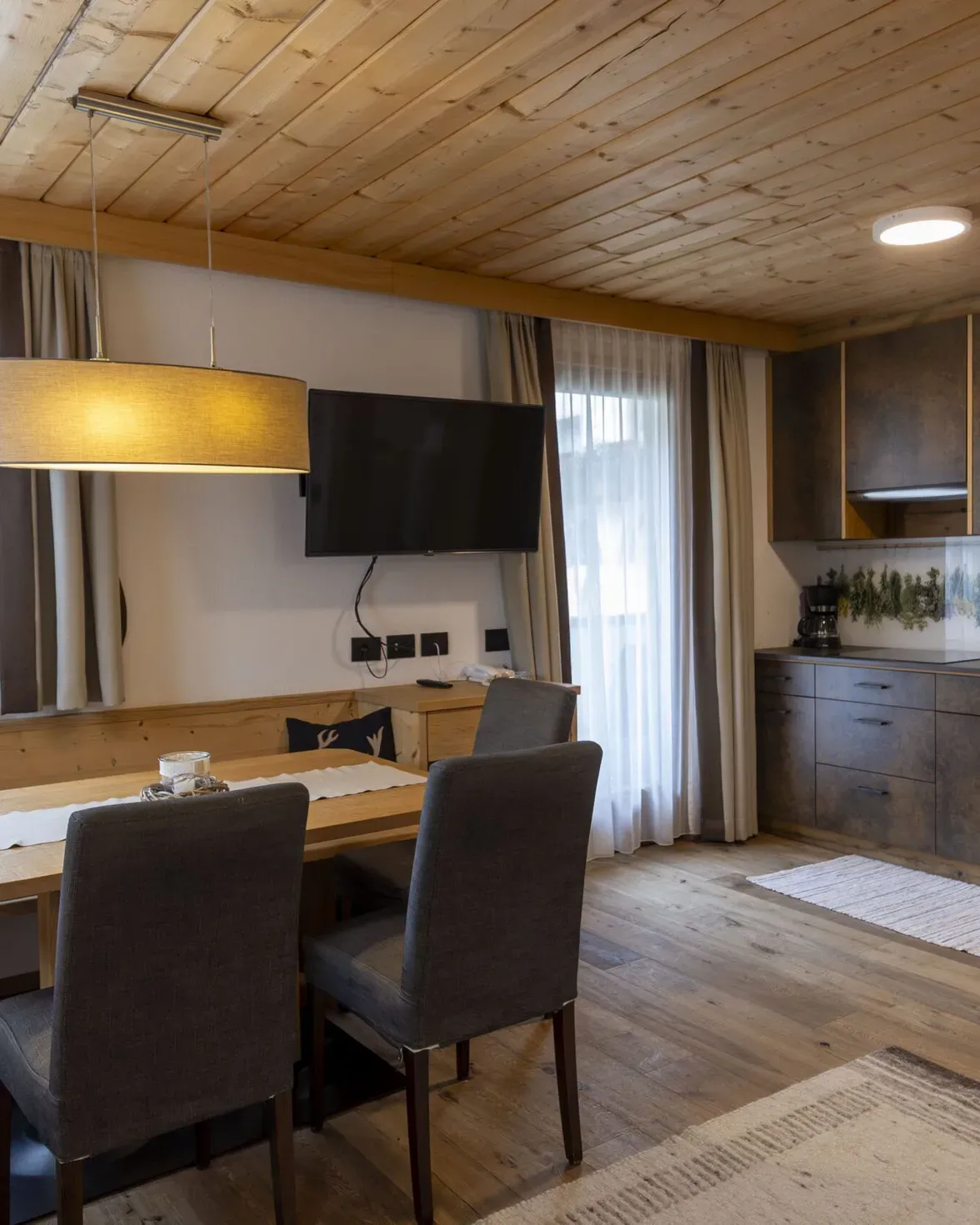 Cozy alpine dining area with wooden ceiling, solid wood table, gray chairs, and a wall-mounted TV.
