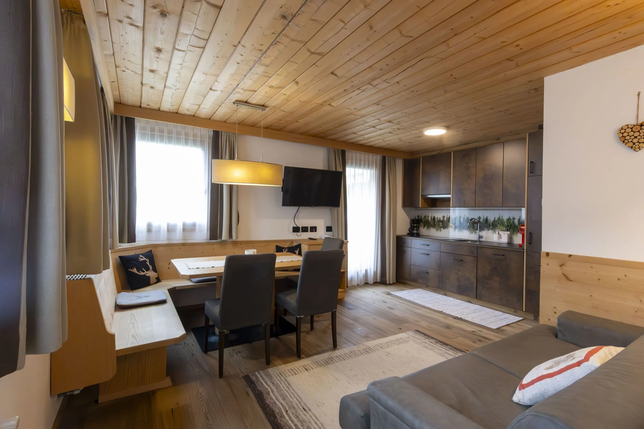 Cozy alpine living room with wooden ceiling, dining nook, wall-mounted TV, and a modern kitchenette.