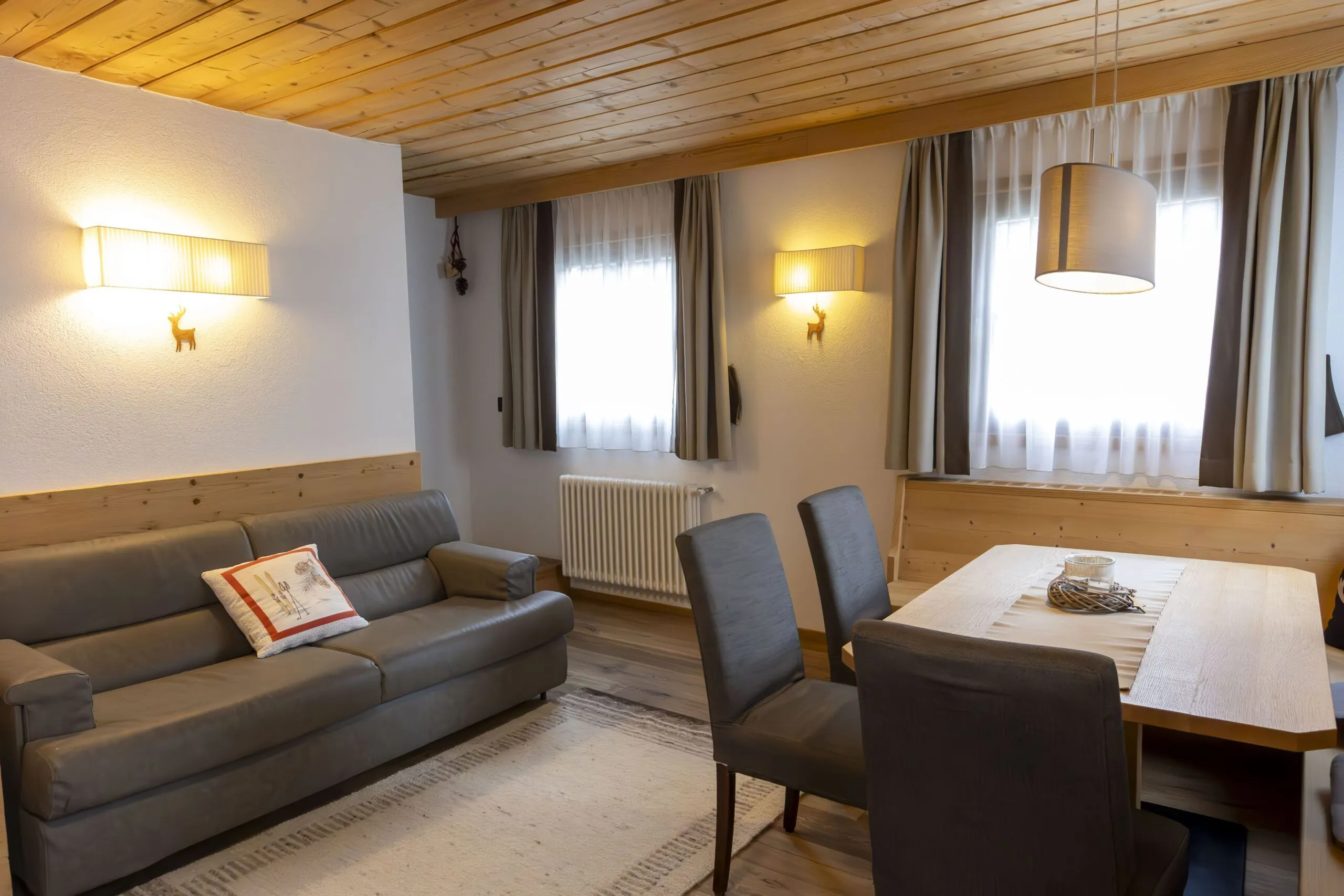 Cozy living and dining area with gray sofa, wooden ceiling, curtains, and a rectangular table.