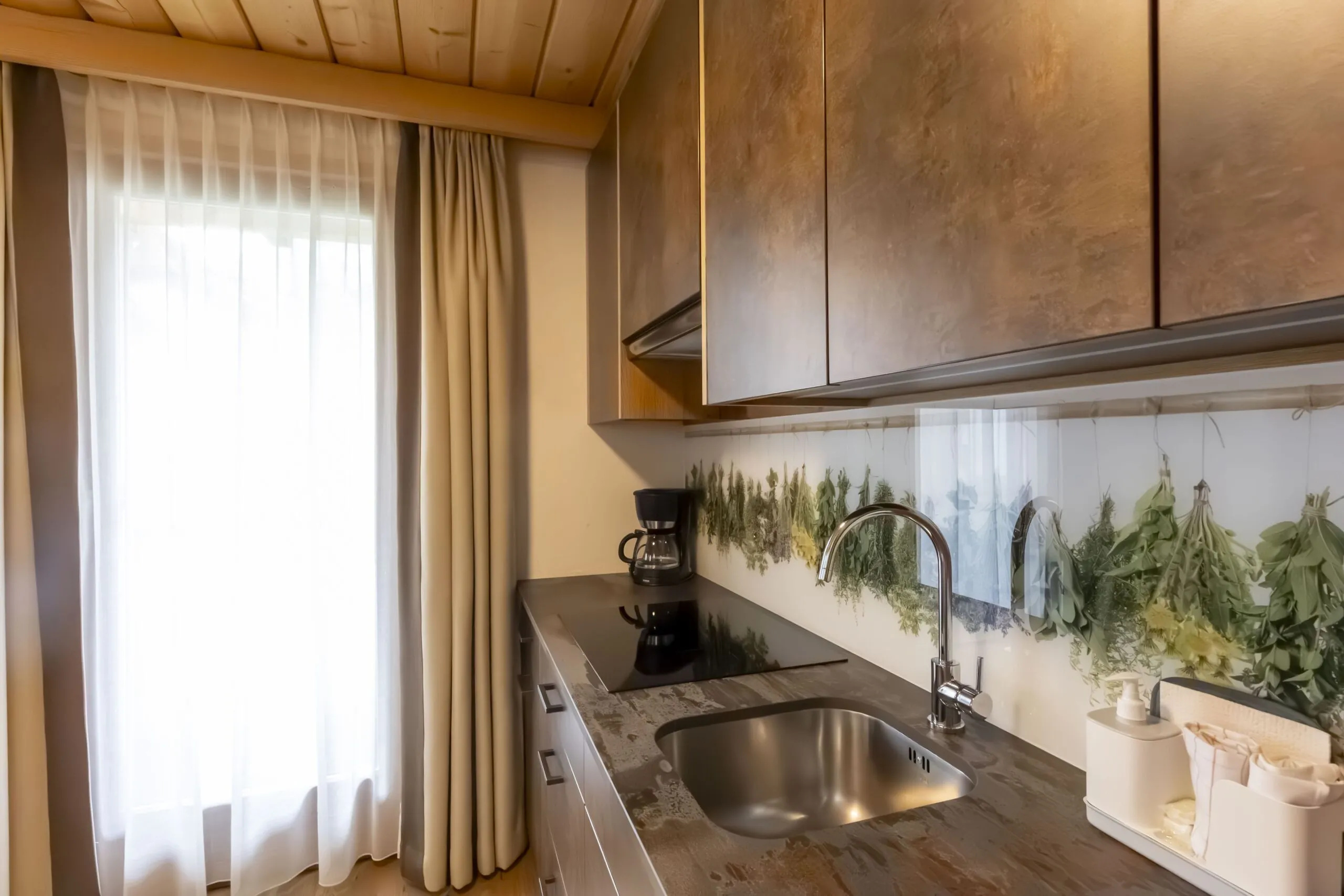 Cozy kitchen with wooden cabinets, beige curtains, and herb-themed backsplash above a stainless sink and stovetop.