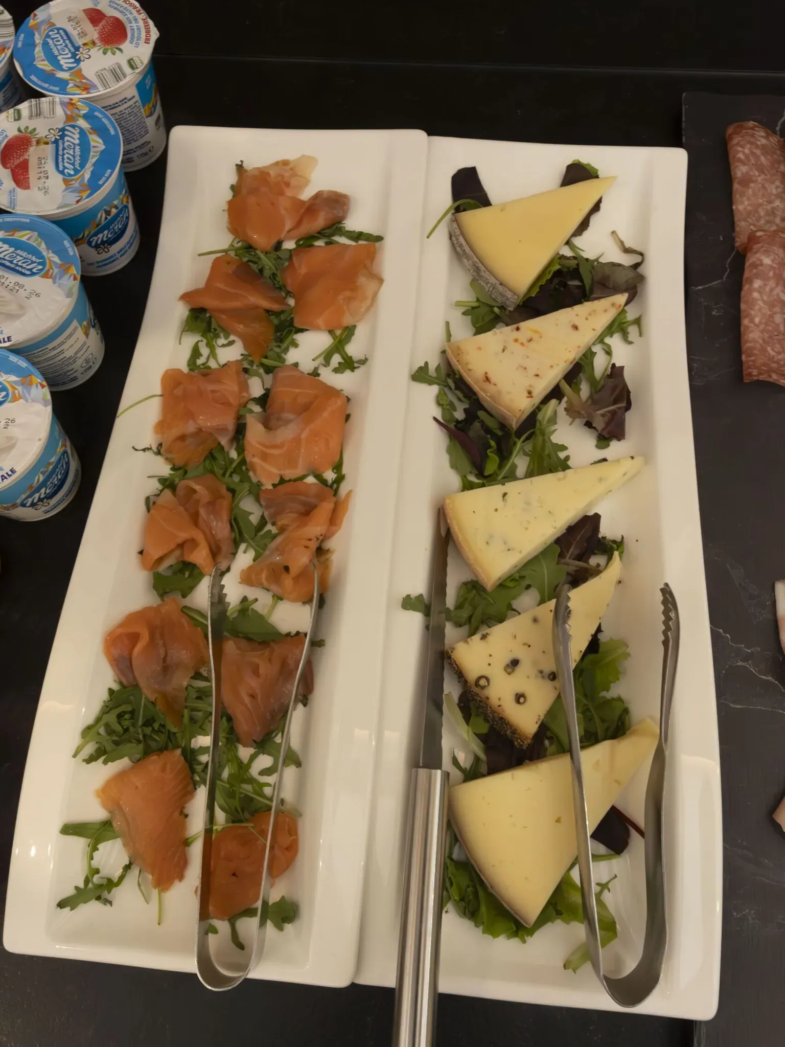 Smoked salmon slices on arugula with white cheese wedges and mixed greens on white platters.