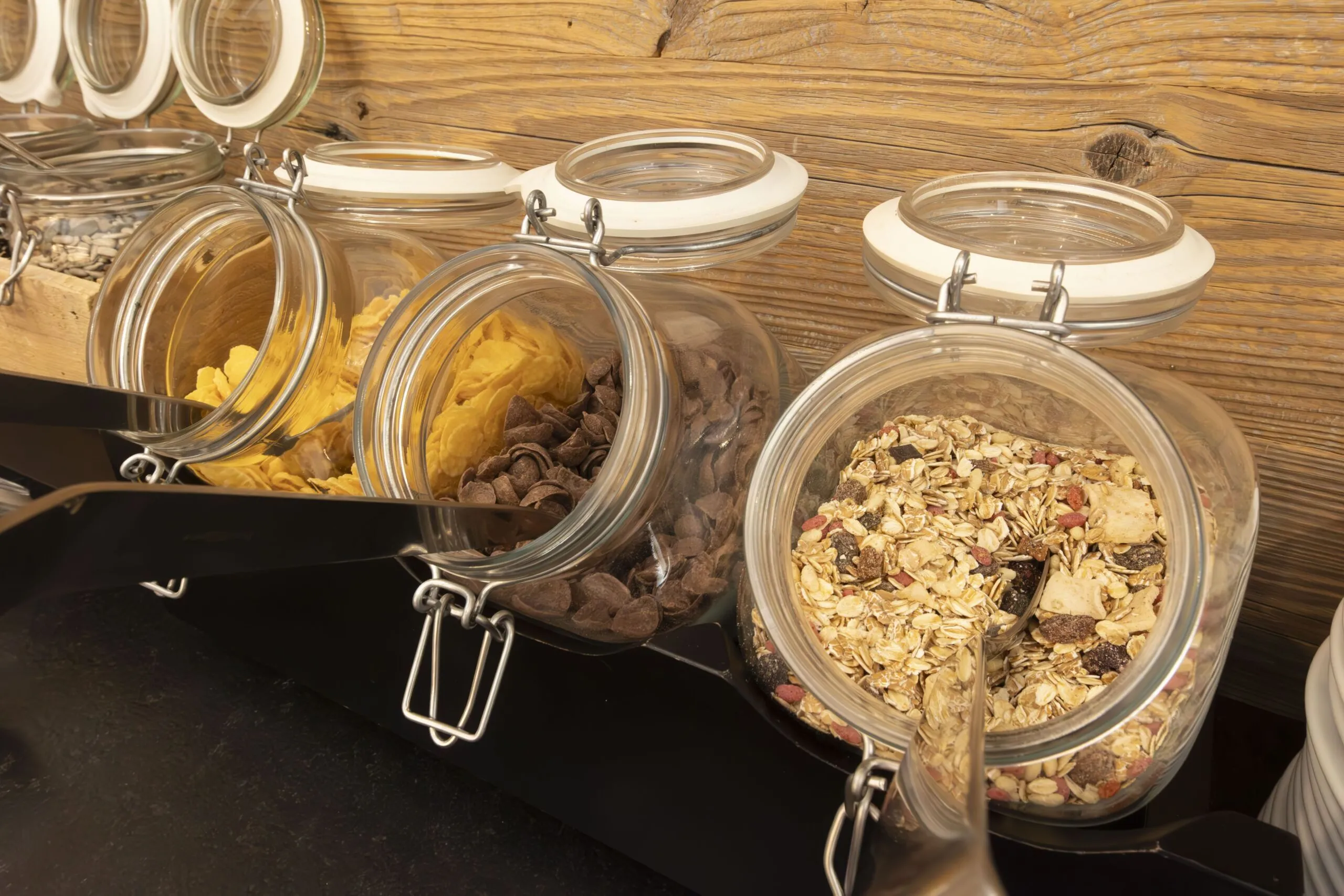 Glass jars with hinged lids on a wooden shelf, filled with cereals, chips, and chocolate chips.