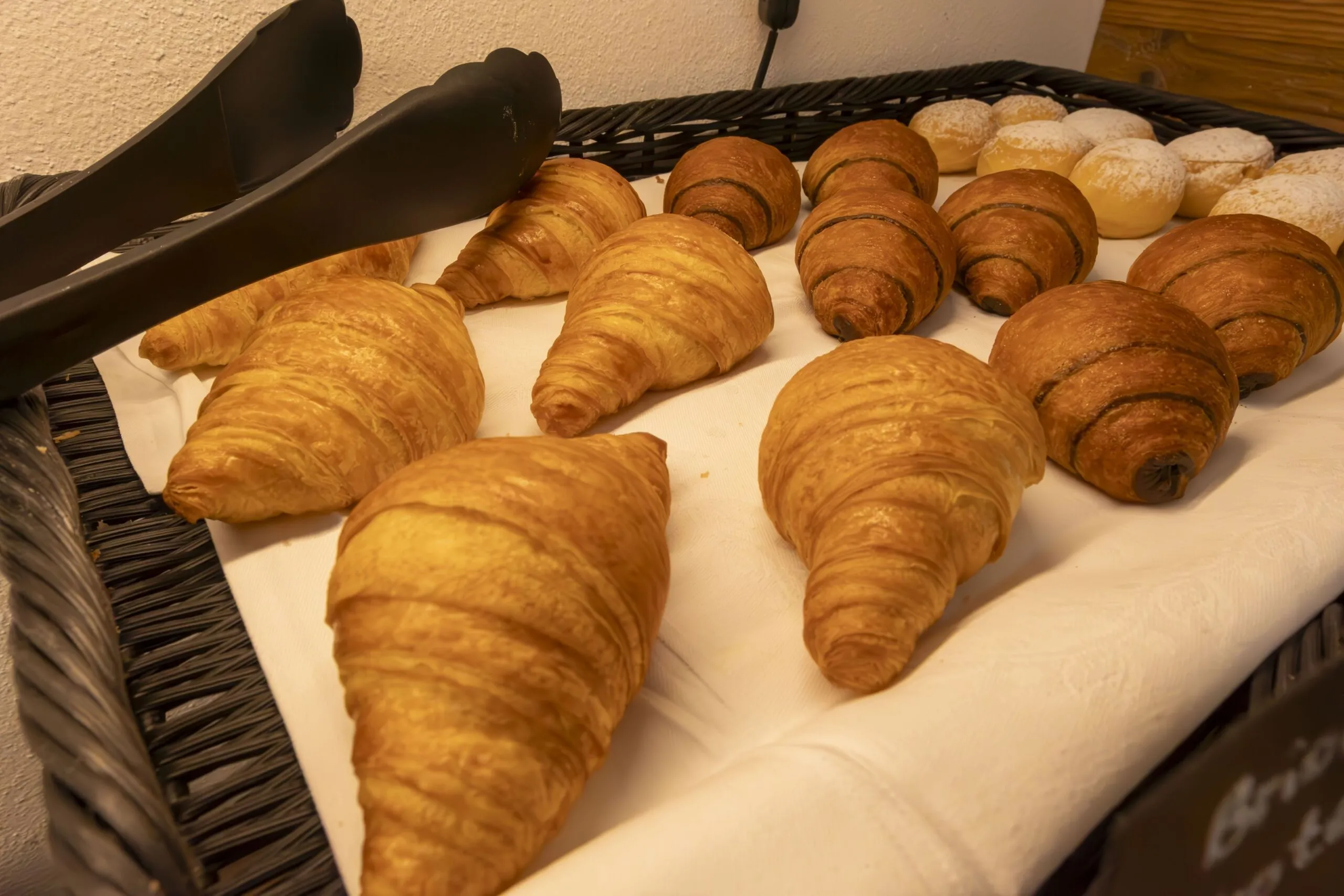 Assorted croissants on a white cloth in a wicker basket, with tongs nearby.