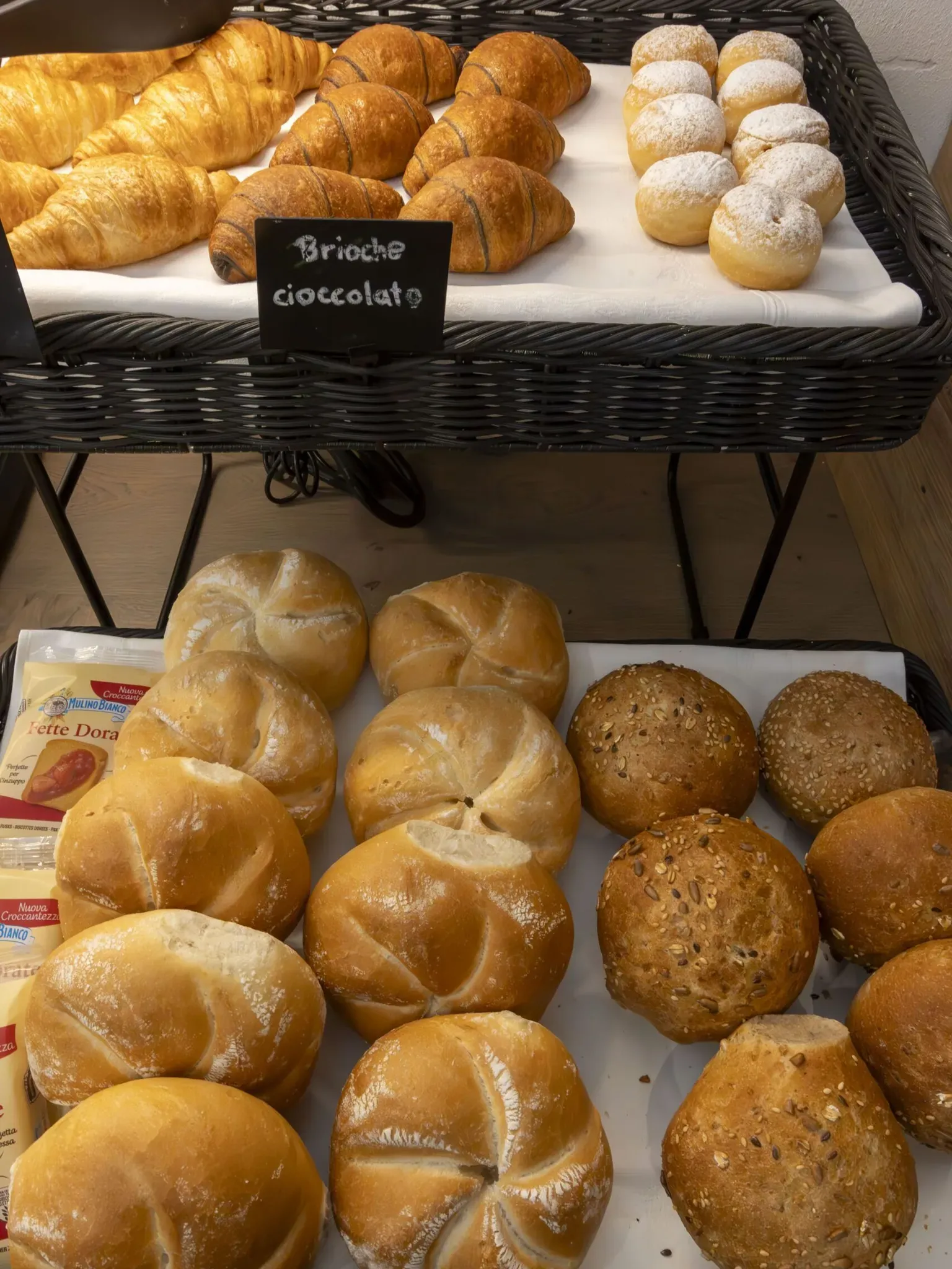 Assorted bread rolls and buns on display, including croissants, round rustic loaves, and sesame sesame-topped rolls in a wicker bakery basket
