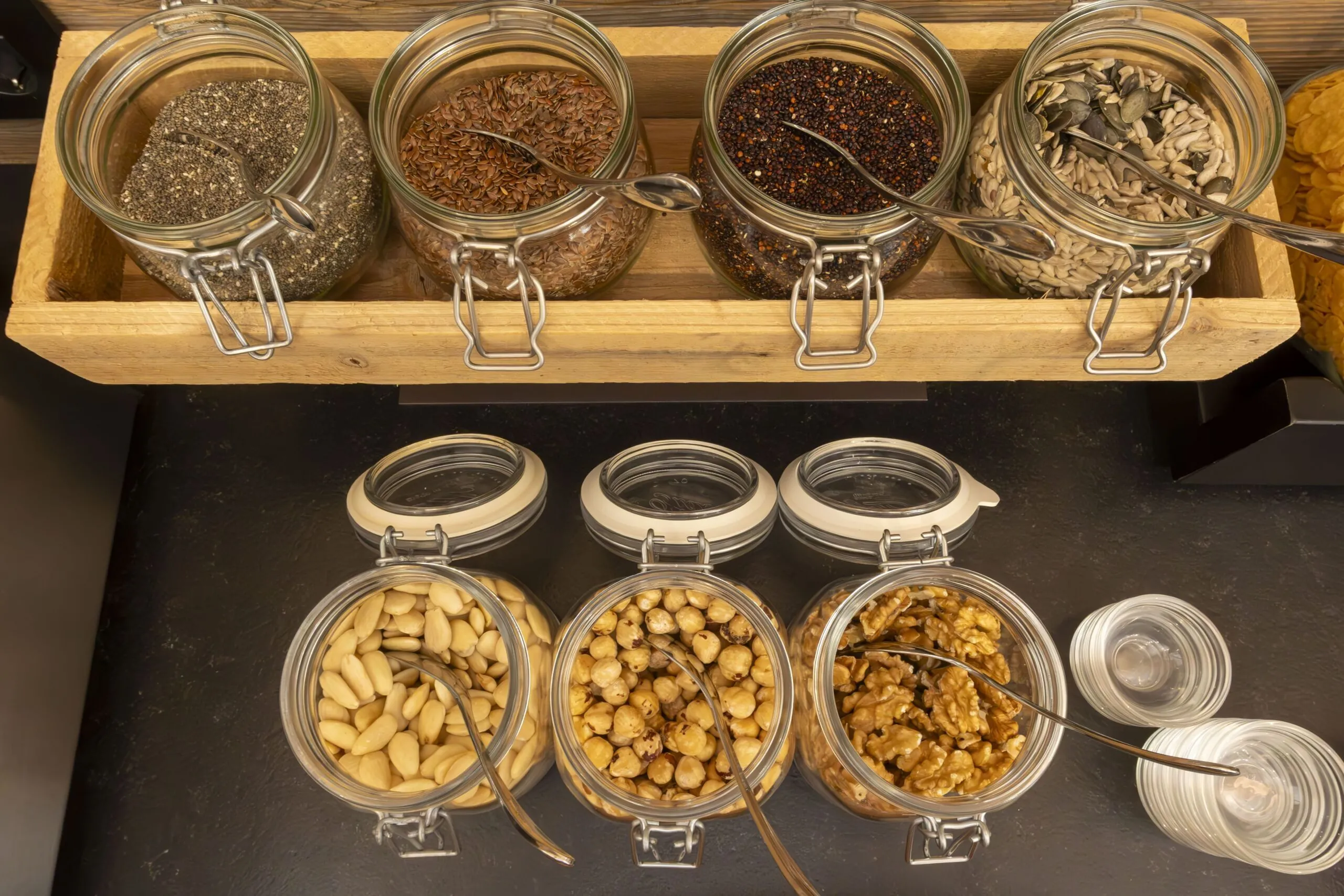 Assorted nuts and seeds in glass jars on wooden shelf, with spoons and lids in view.