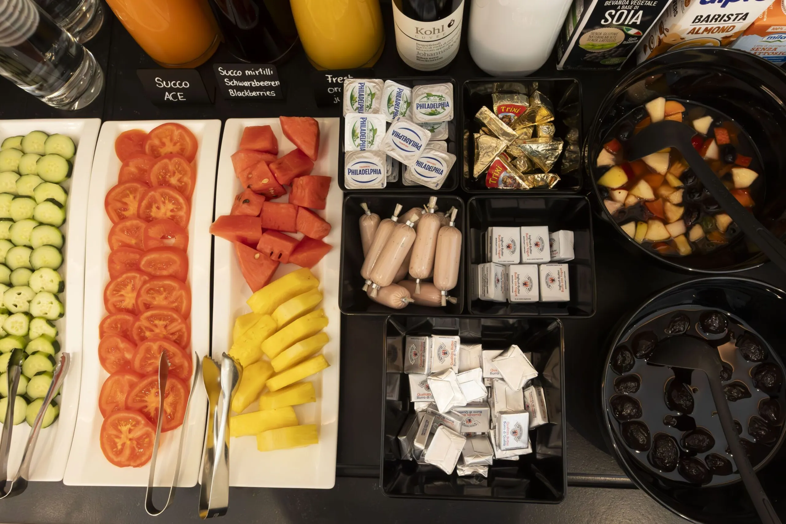 Assorted fruit and toppings in a self-serve bar: cucumber slices, tomato rounds, watermelon chunks, pineapple strips, cheeses, sausages, and bowls of fruit salad.