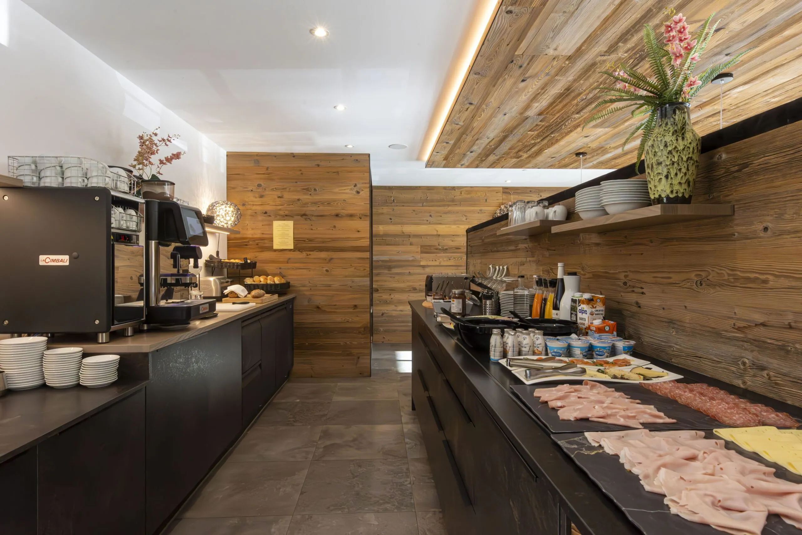 Warm wood-toned cafe counter with coffee machine, pastries, and a deli spread on a dark countertop.