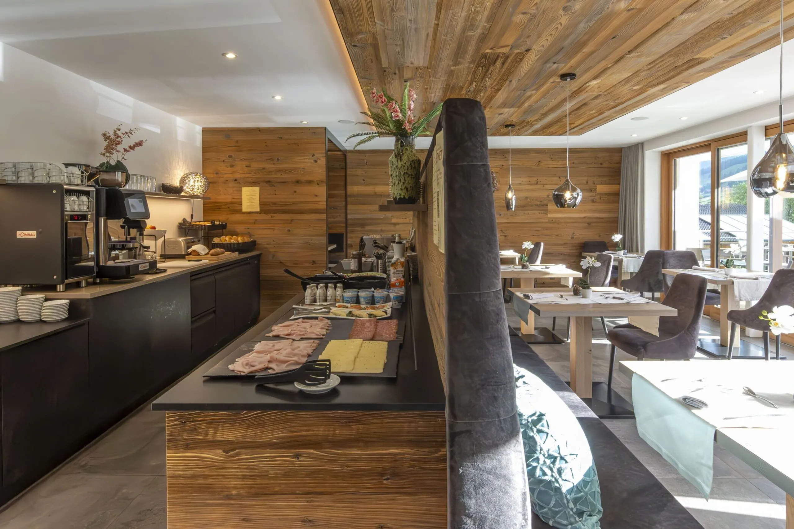 Hotel breakfast area with a long dark counter, coffee station, bread display, and wooden walls; bright seating by large windows.