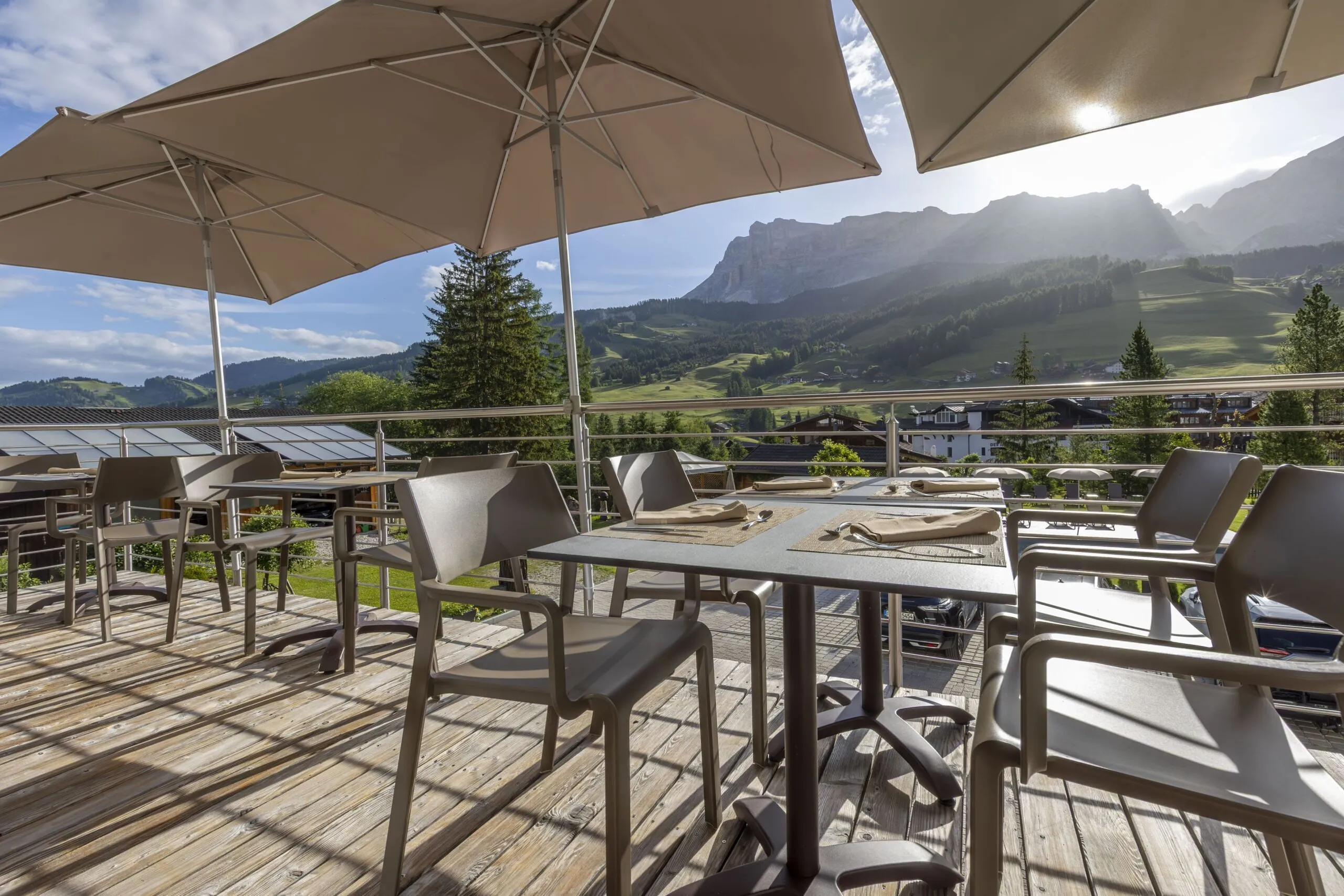 Outdoor restaurant terrace with tables and chairs under large umbrellas, overlooking green hills and mountains in Alta Badia.