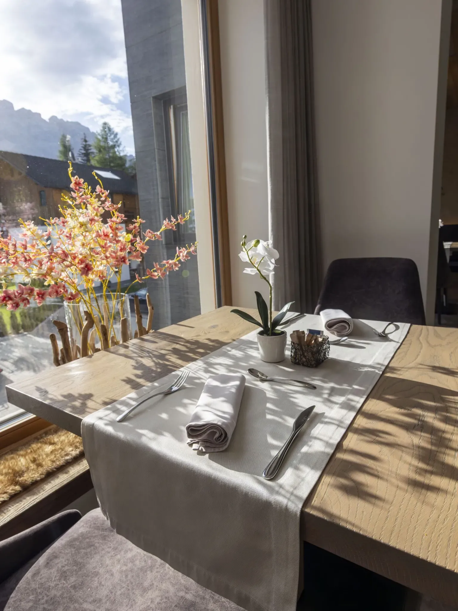Table set by a window with a vase of pink blossoms, white orchid, and sunlight casting shadows.