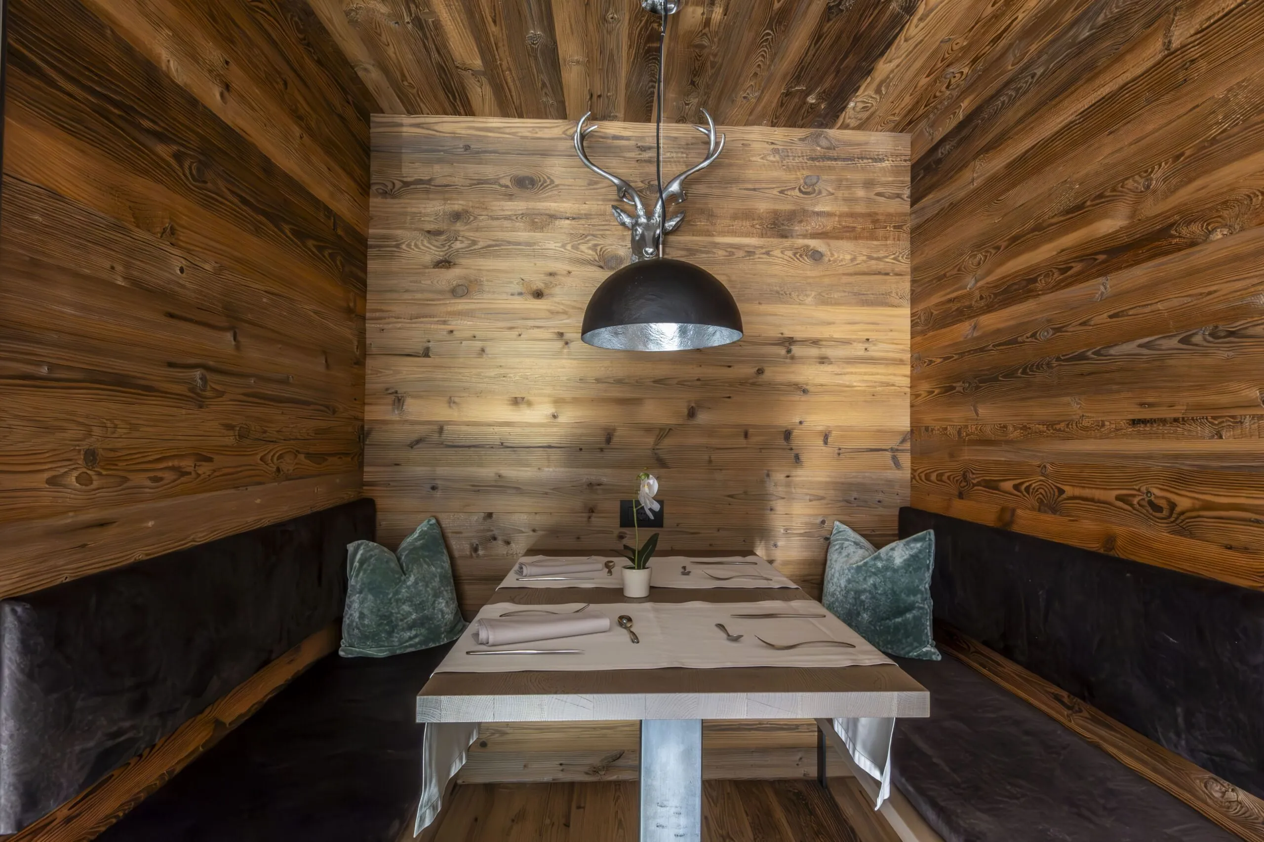 Cozy wooden dining nook with a central table, deer-horn pendant light, and velvet pillows.