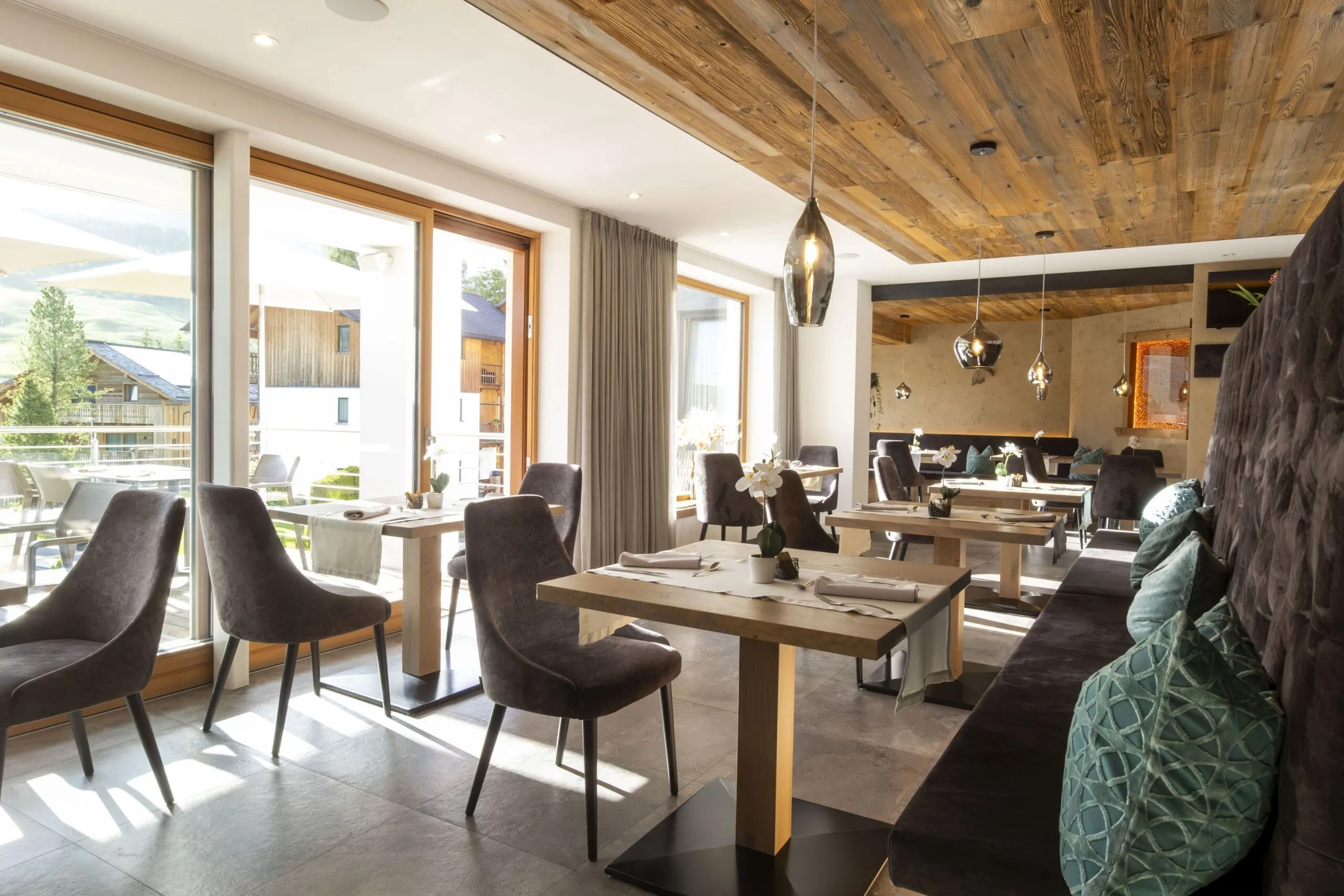 Restaurant dining area with wooden ceiling, large windows, grey chairs, and light wooden tables.