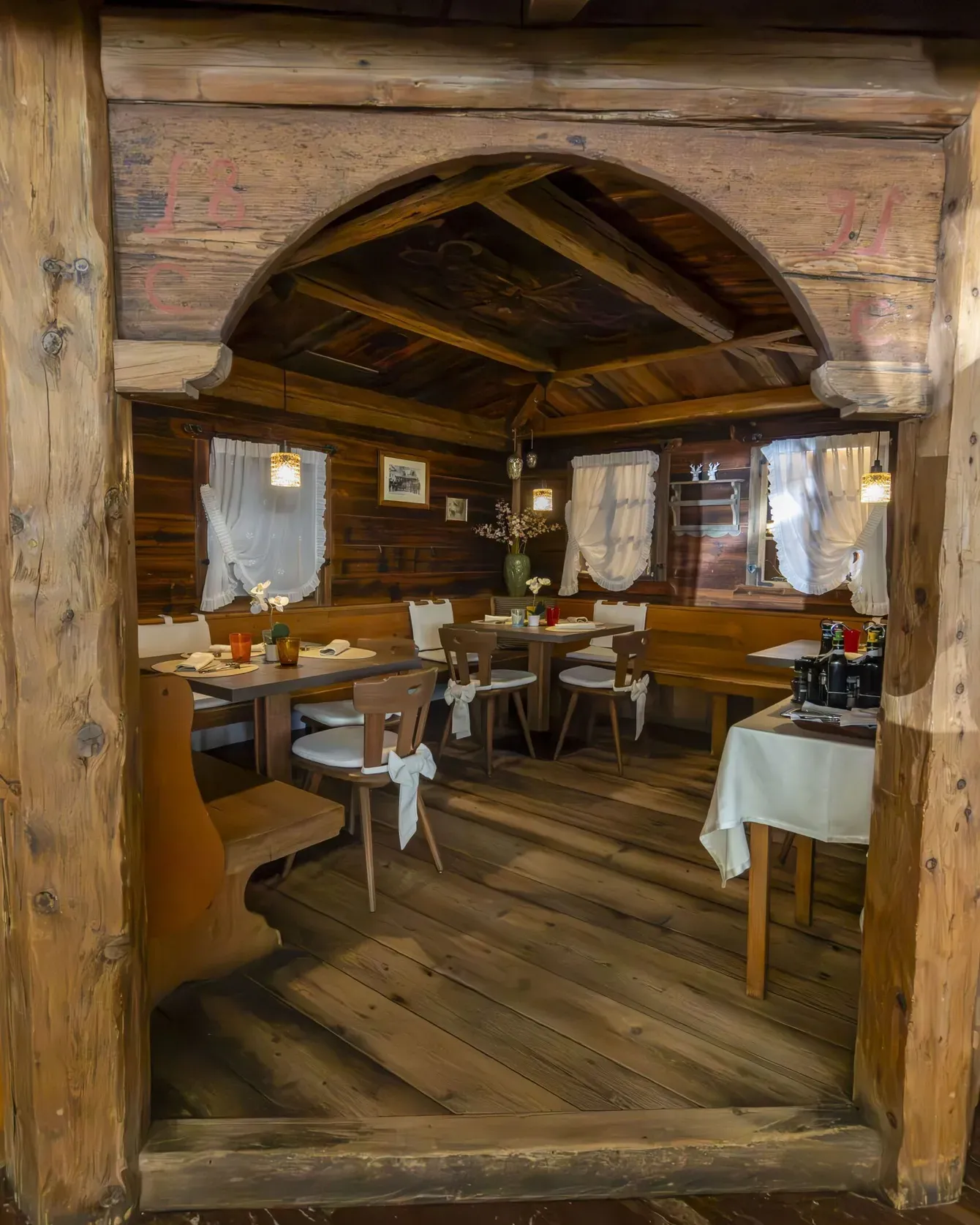 Cozy rustic dining room in a wooden lodge with white curtains, carved beams, and tables set for meals.