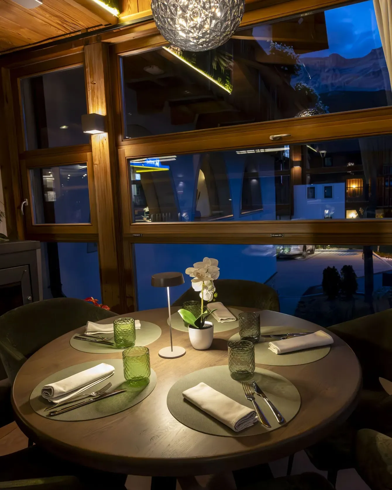 Cozy restaurant table set for four by large wooden windows with a glowing pendant light and orchid centerpiece at night