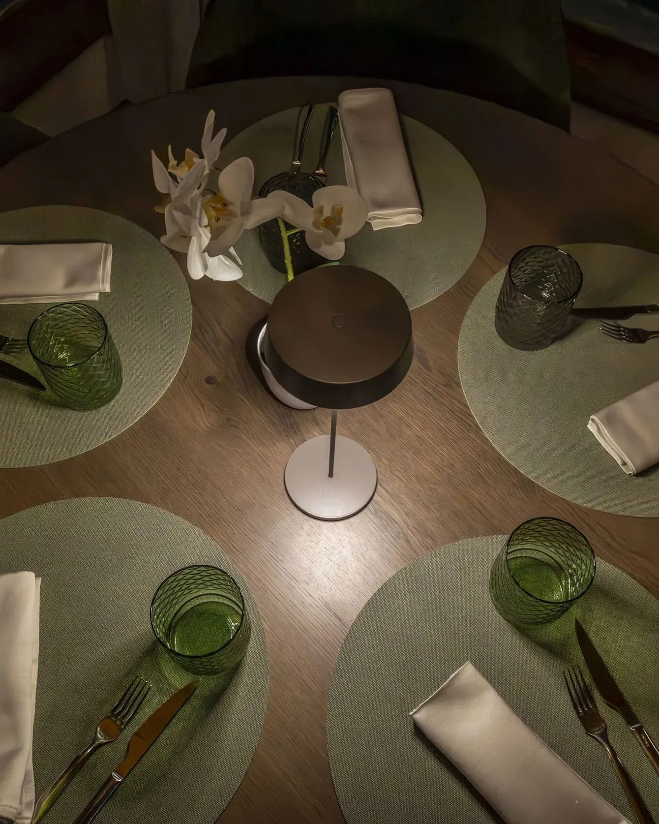 Dining table setup with green placemats, white napkins, cutlery, green glassware, and a central flower arrangement.