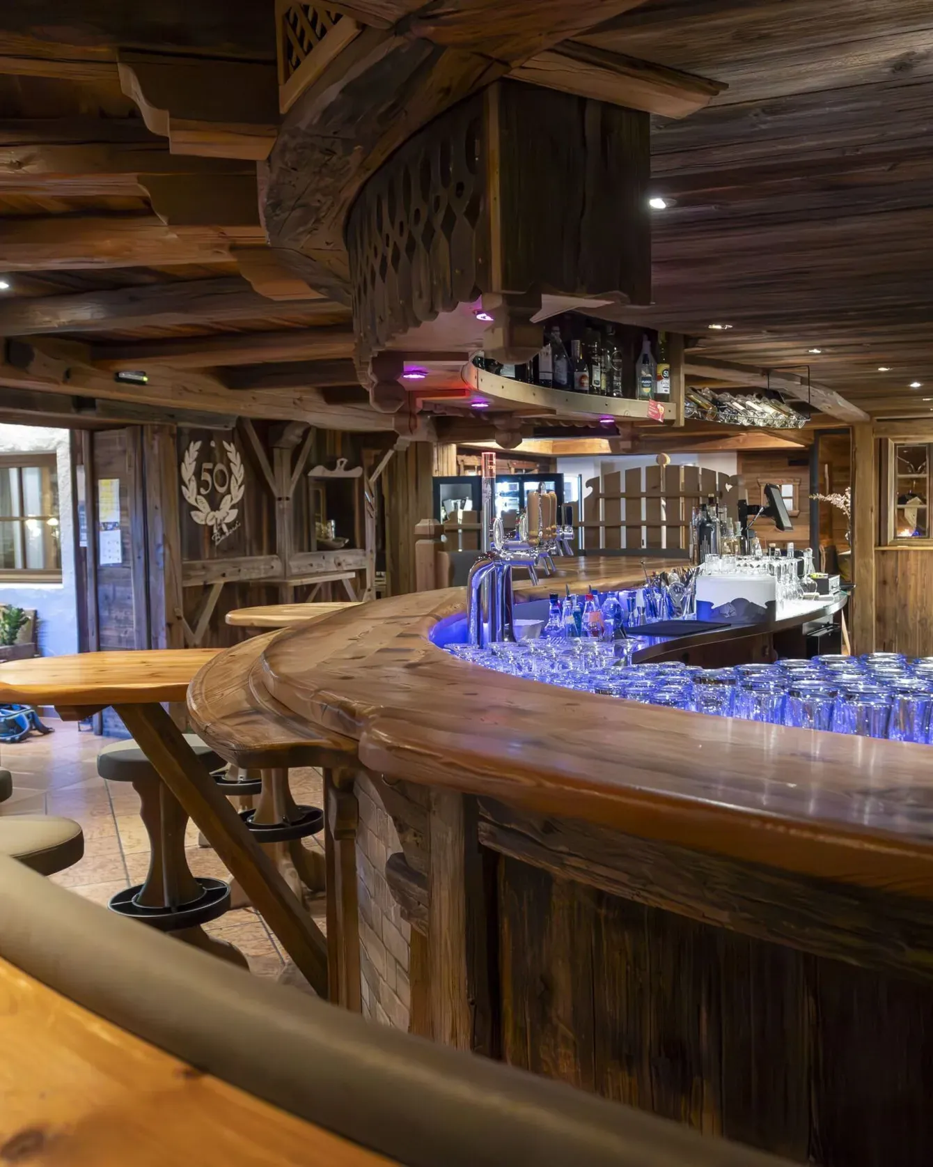 Cozy wooden bar interior with curved counter, blue lighting, bottles, and stools in rustic tavern.