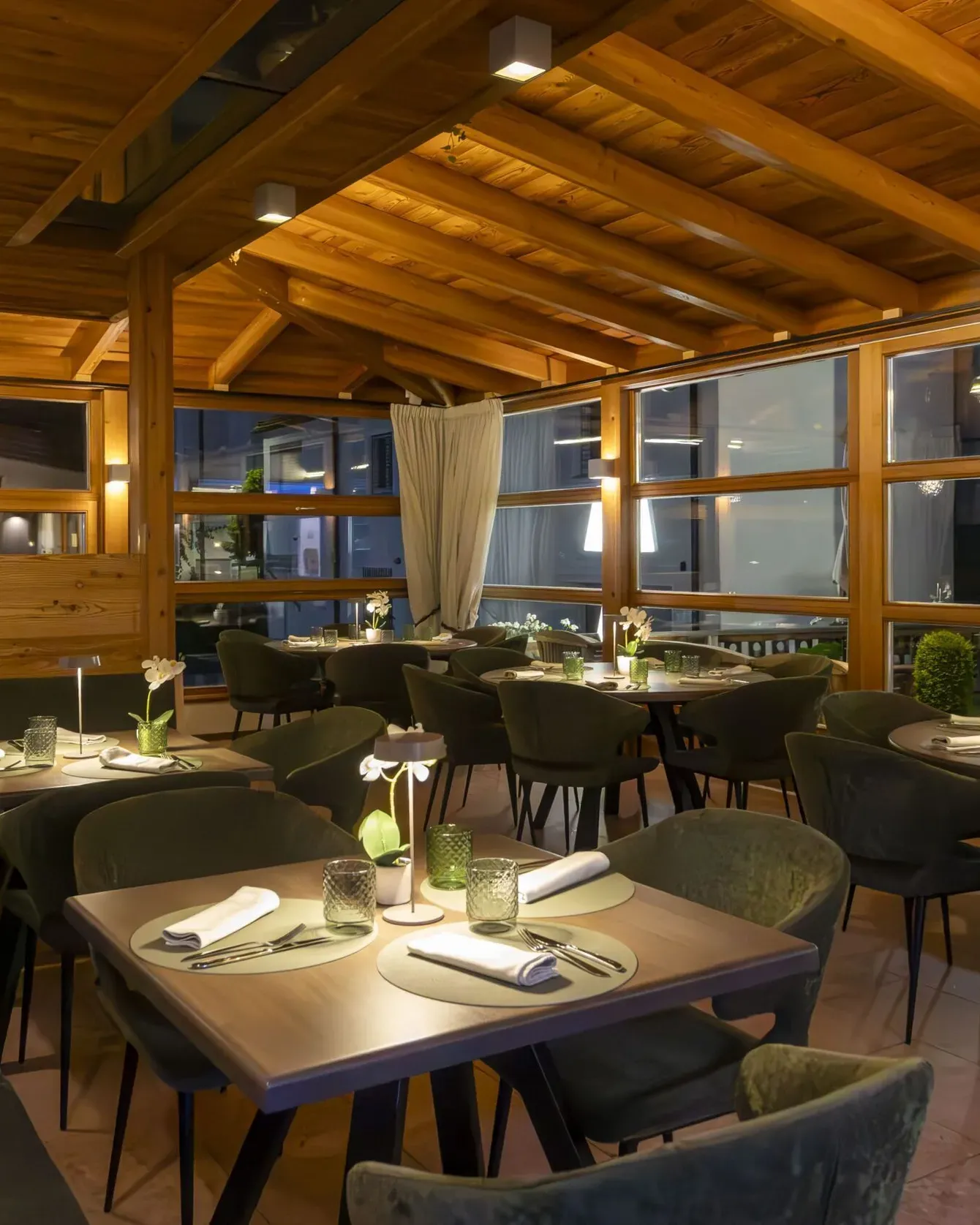 Cozy wooden interior restaurant with green upholstered chairs, round tables set for dining, warm lighting, and large windows.