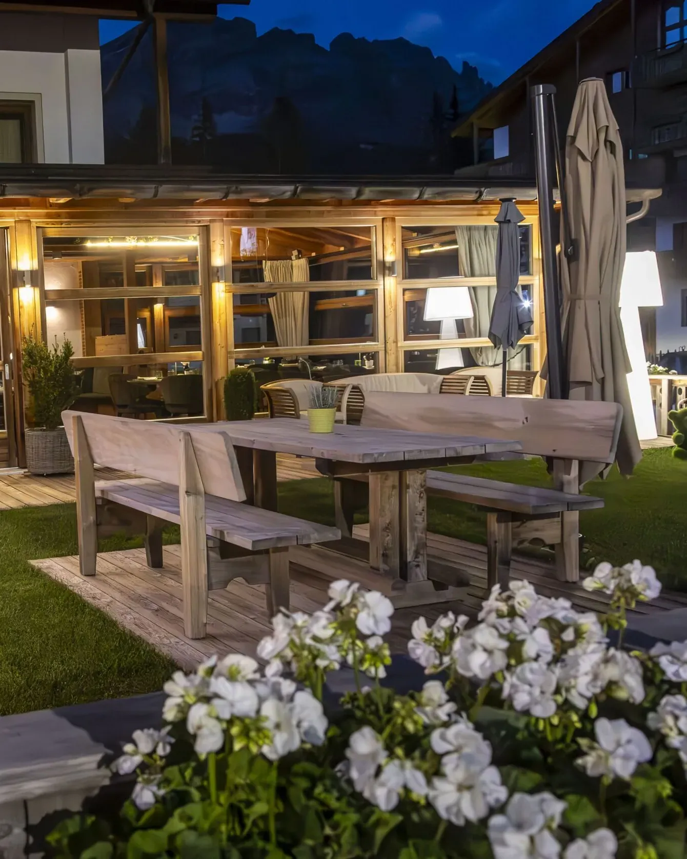 Night-time outdoor dining area with wooden tables, bench seating, and glass-walled restaurant interior in Alta Badia.