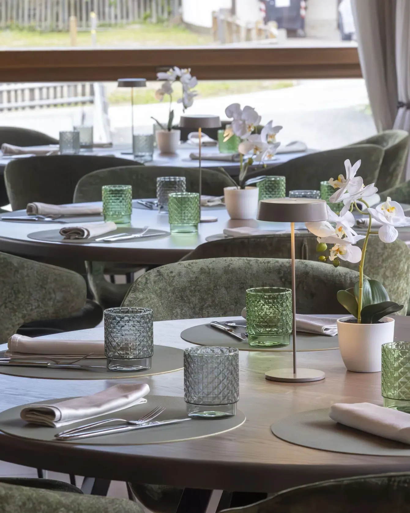 Elegant restaurant interior with round tables, green velvet chairs, and orchids in soft lighting.