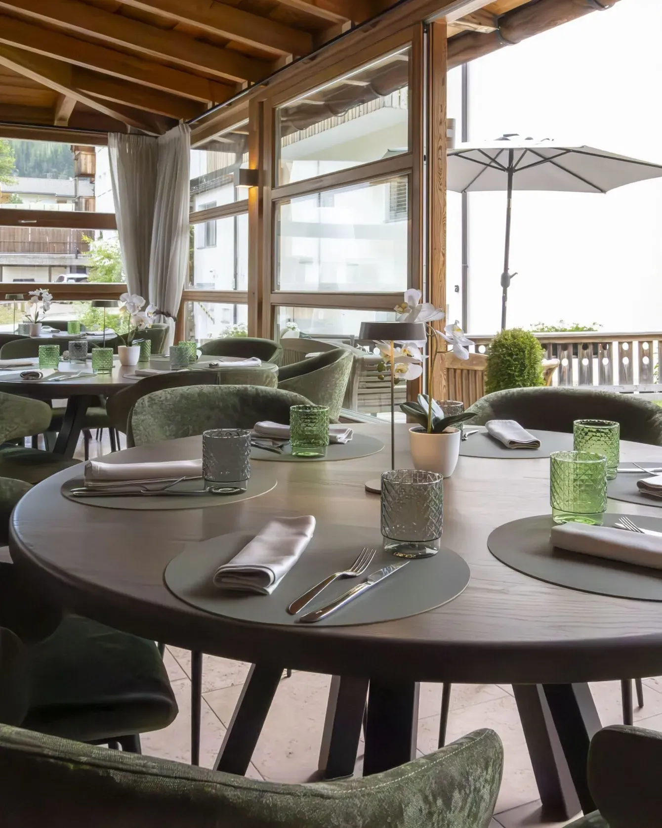 Elegant dining area with round tables set for guests, green upholstered chairs, and a wooden screened veranda.
