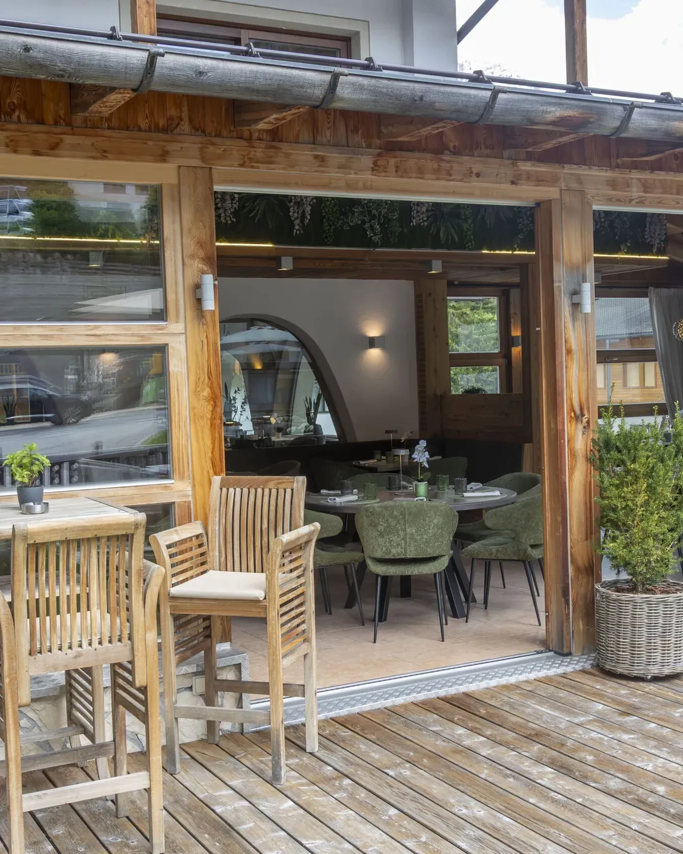 Wooden patio dining area with tables and chairs outside a glass-front café, alpine village buildings nearby.