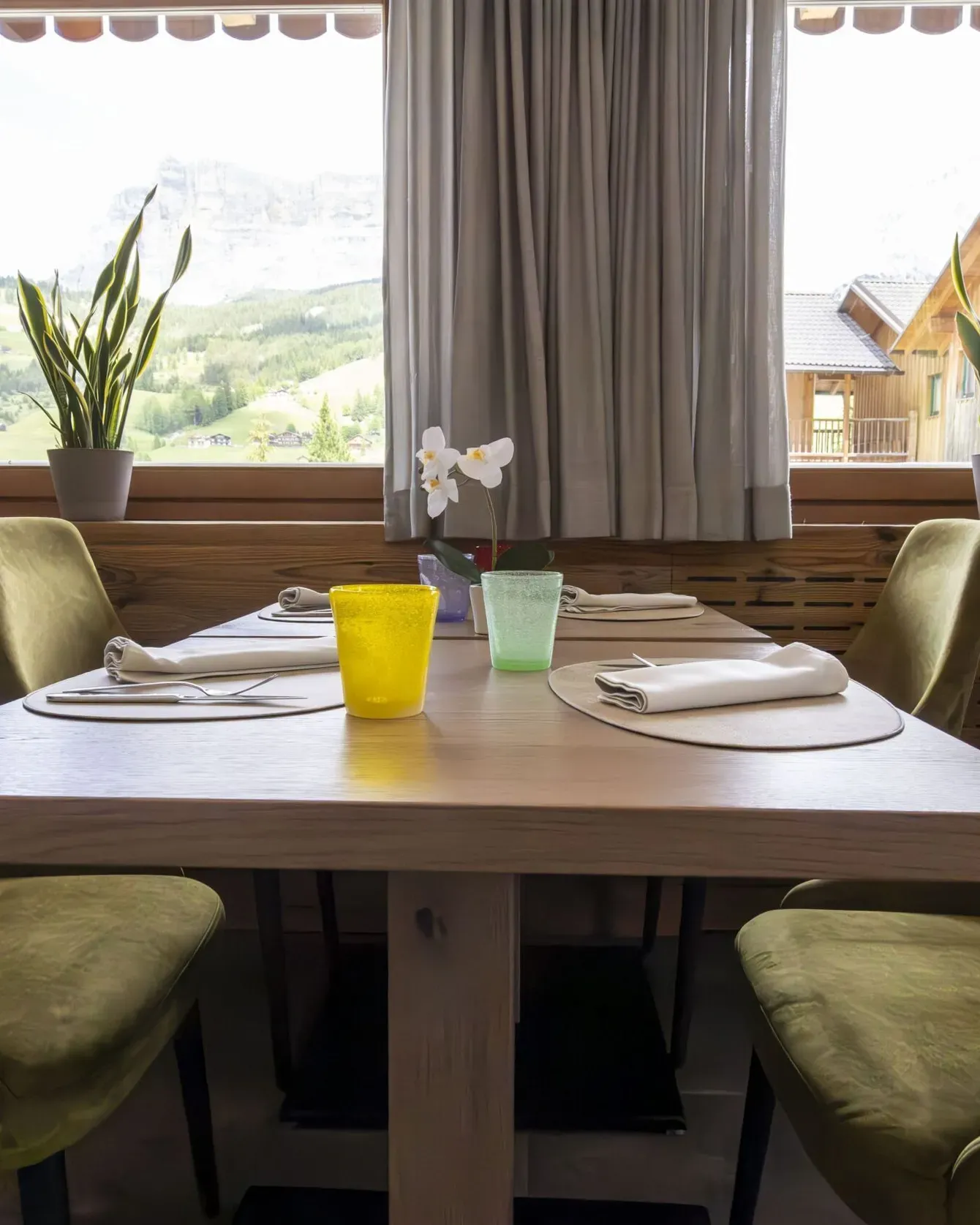 Cozy dining table set for four by a window with green upholstered chairs and potted plants.