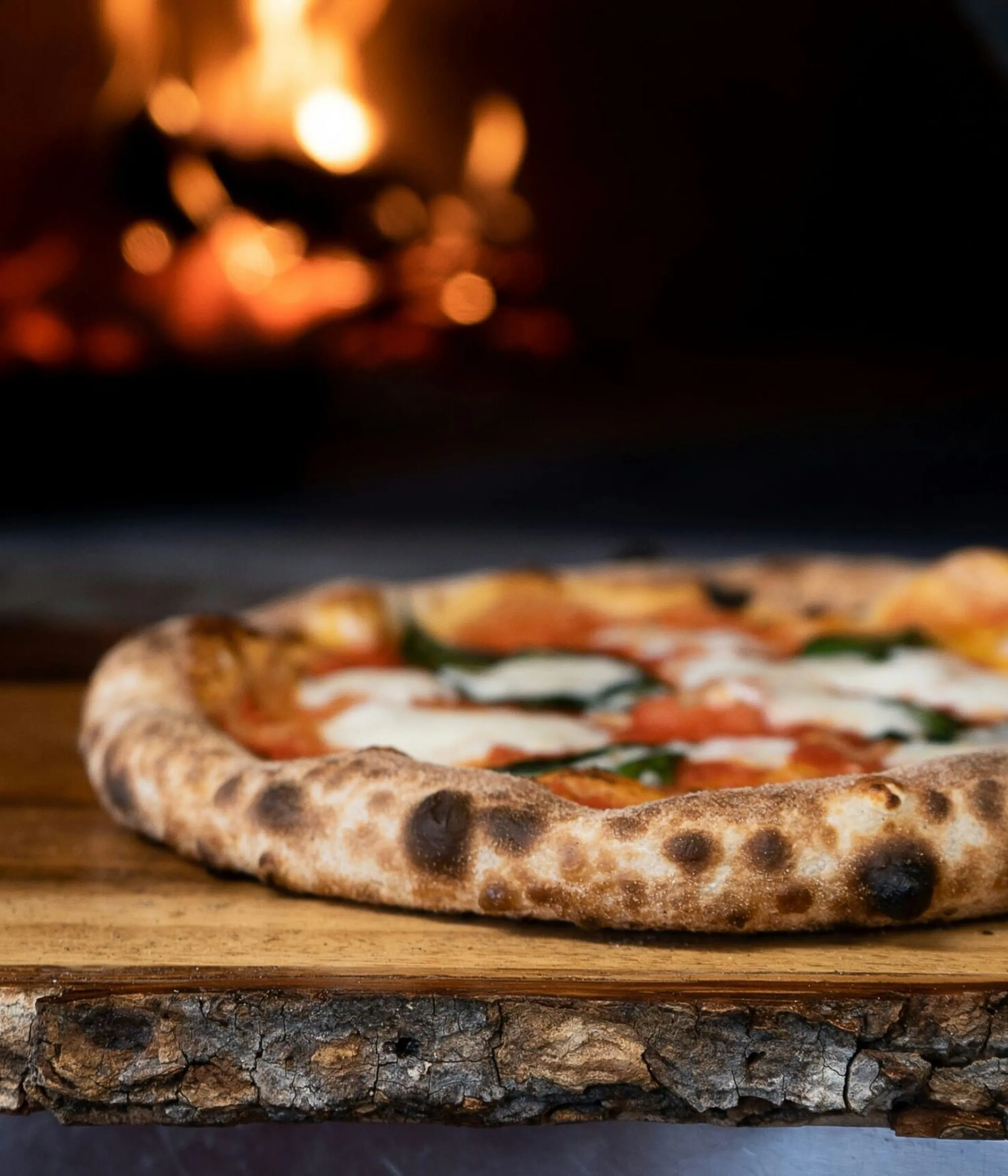 Wood-fired pizza on a rustic wooden table with a glowing oven in the background, charred crust edges visible.