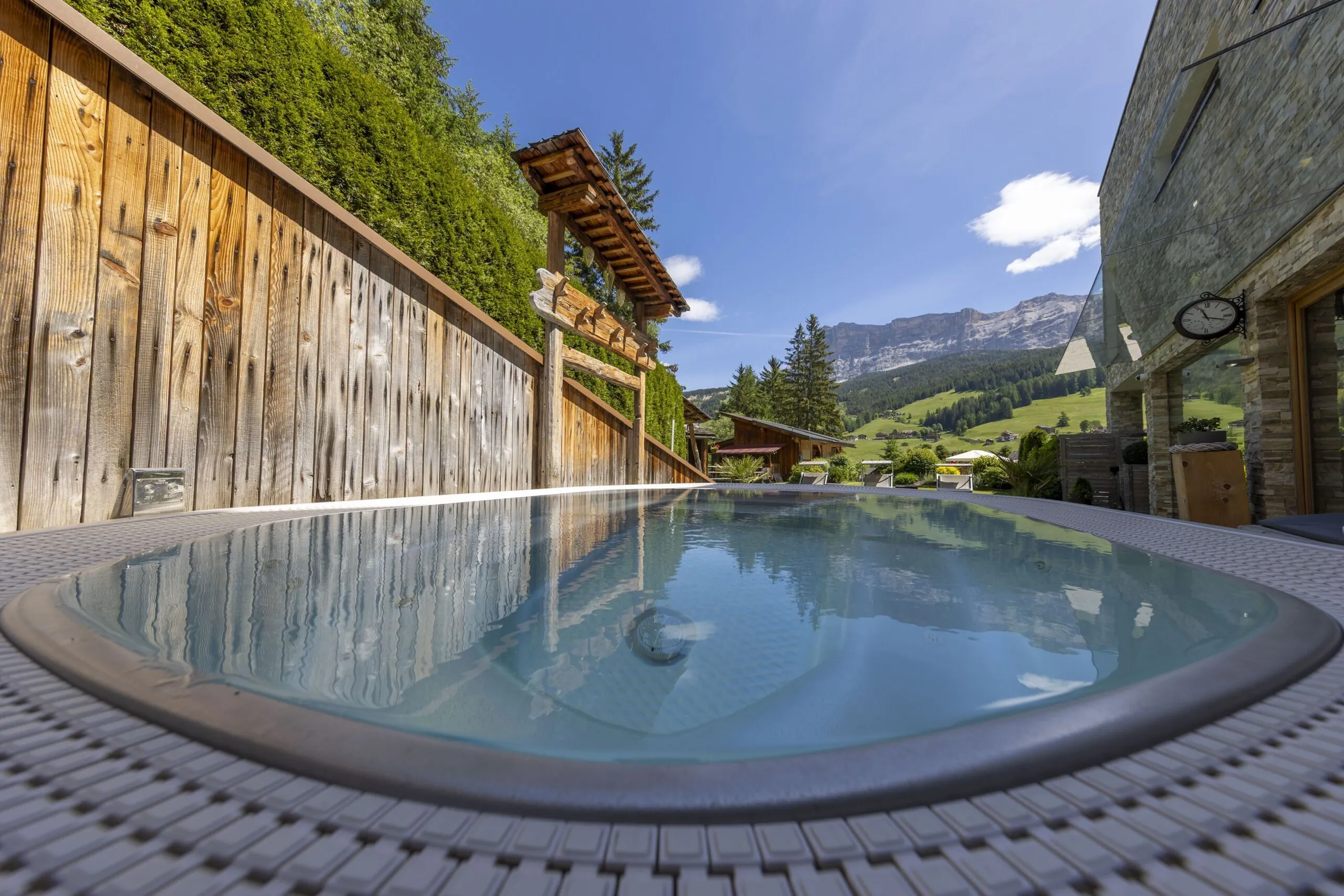 Outdoor hot tub with glassy water, wooden fence, and alpine mountains in the background.