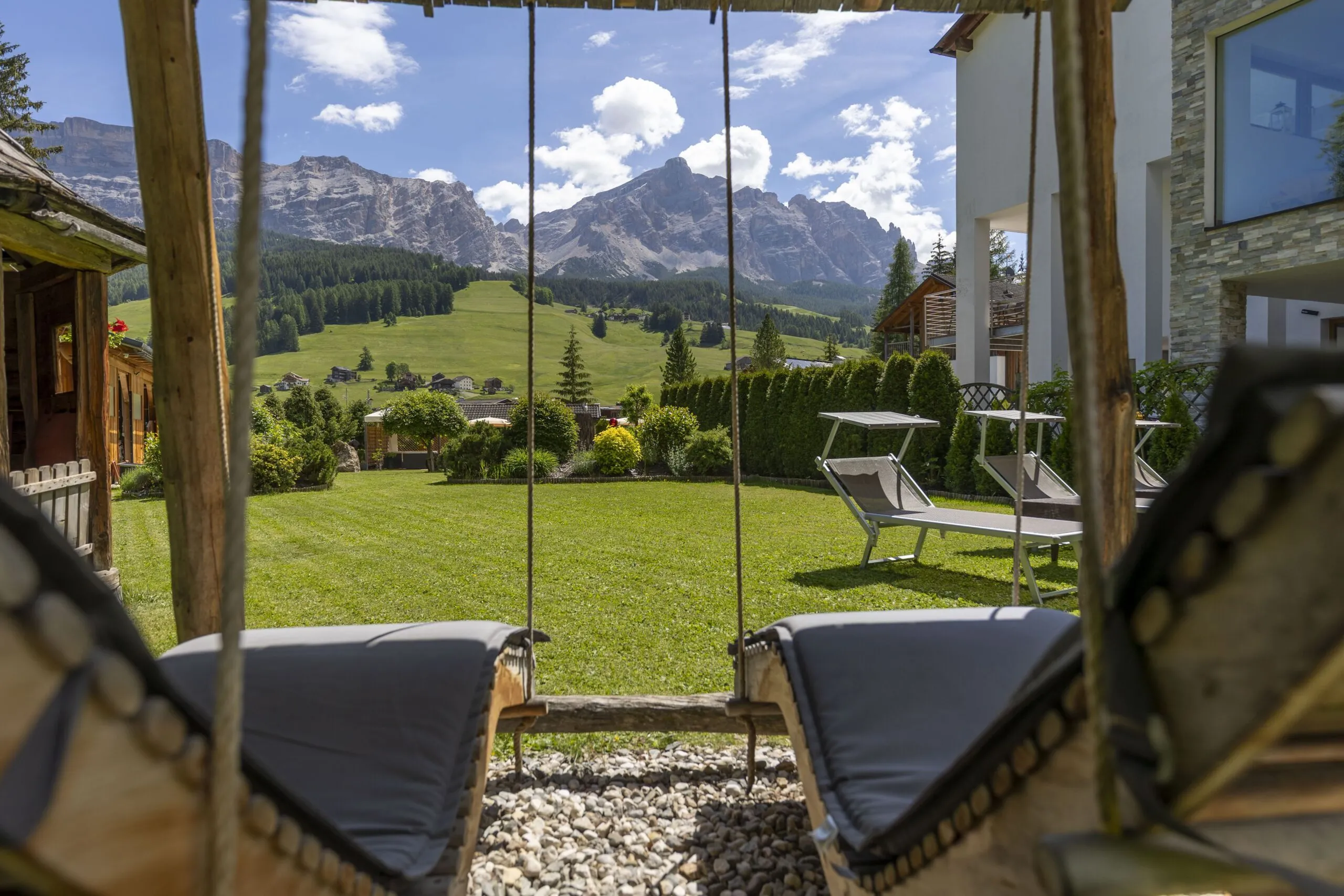 Two wooden swing chairs overlook a green lawn with mountains in the distance under a bright blue sky.