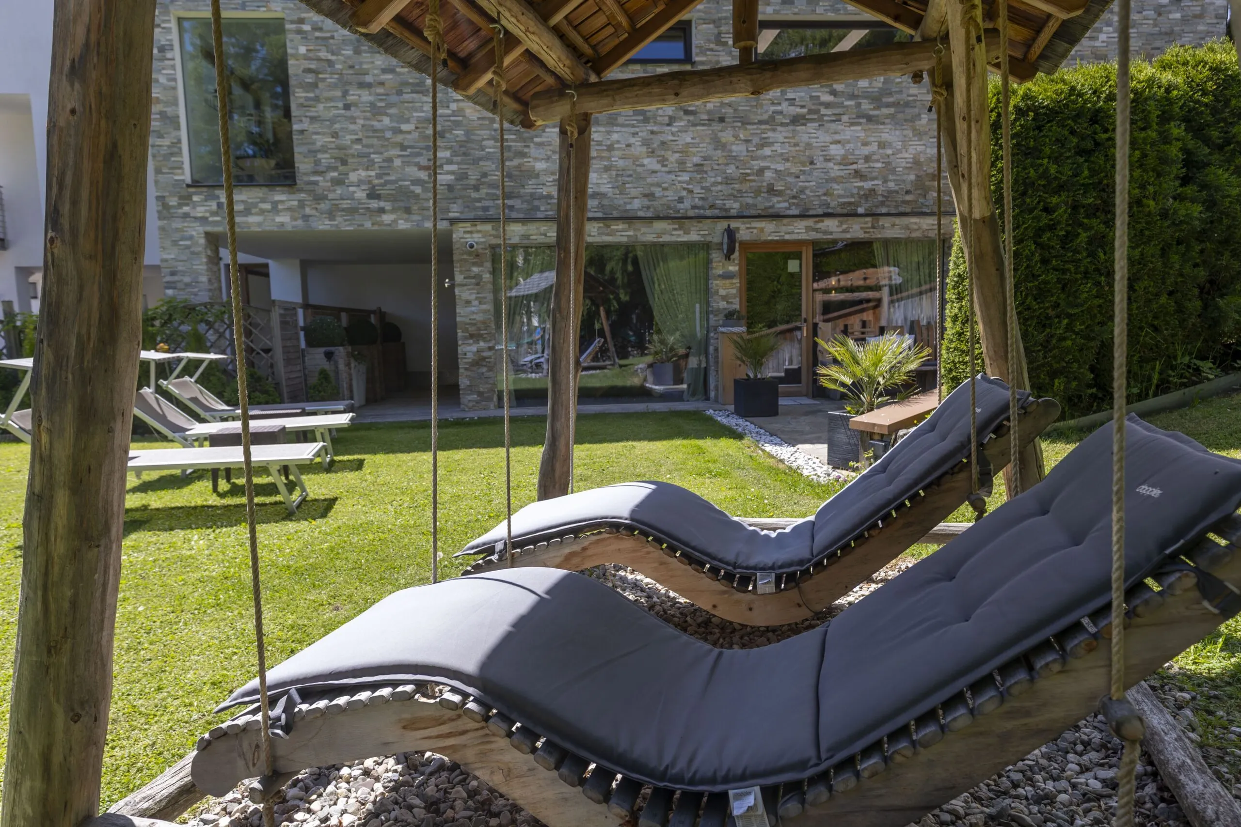 Wooden outdoor swing with cushions in a yard, modern stone house backdrop and lounge chairs nearby.
