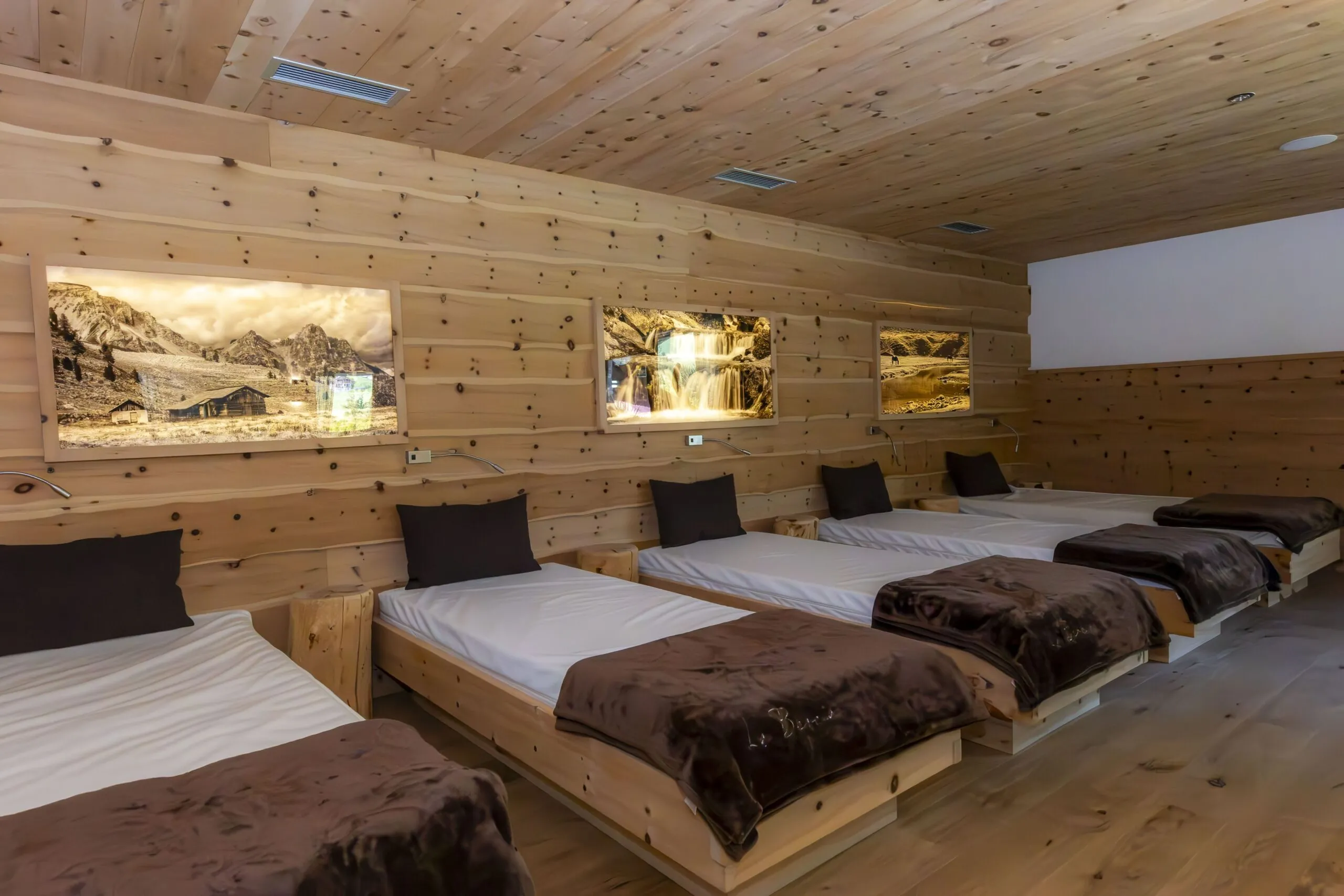Multiple wooden-bed dorm room with white sheets, brown blankets, black pillows, and framed mountain photos on pine walls.