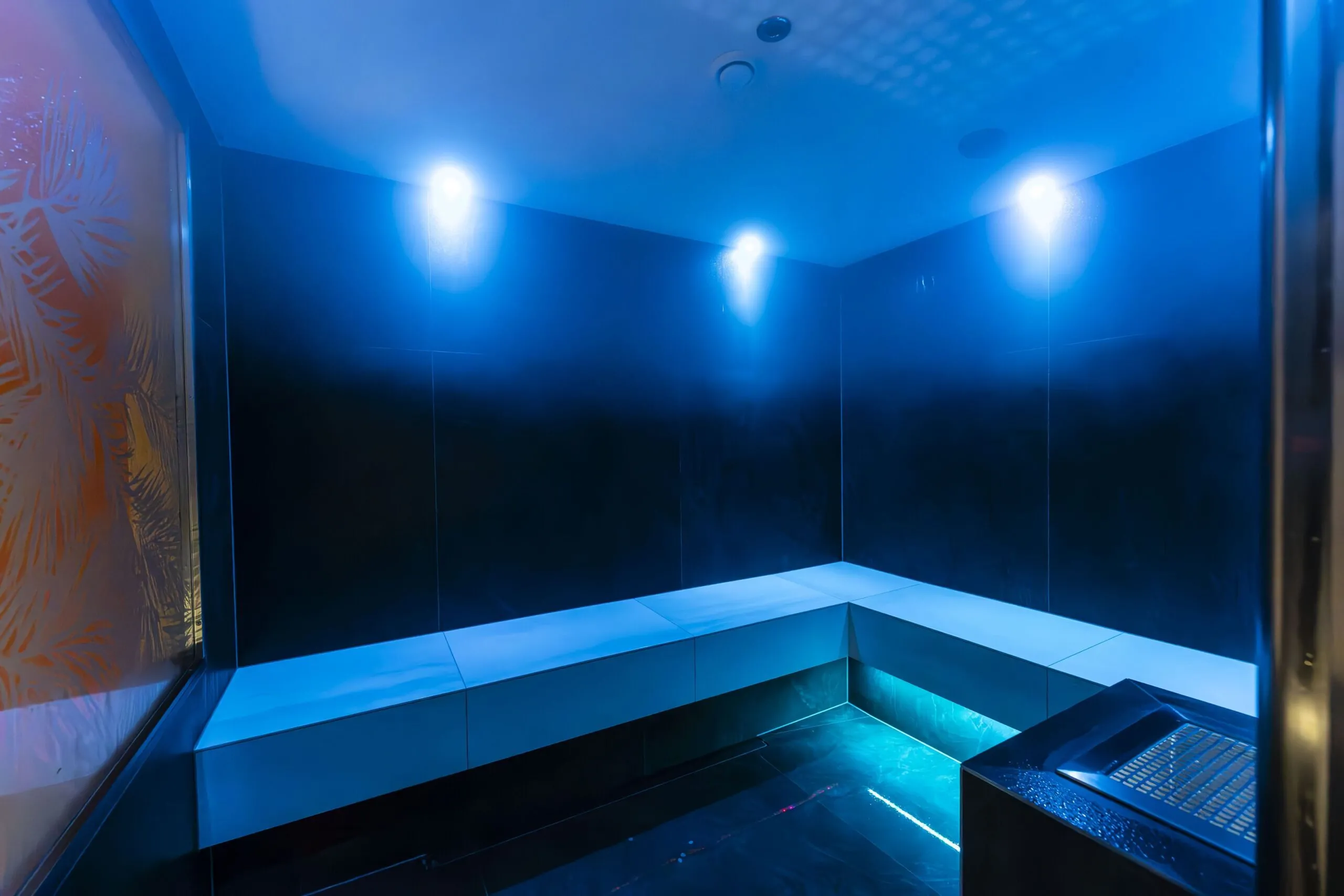 Dark, blue-tinted steam room with corner seating and bright wall-mounted lights.