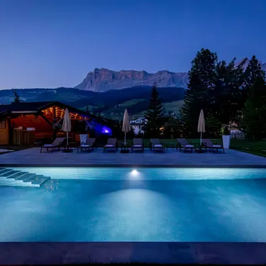 Evening infinity pool scene at a resort with lounge chairs, umbrellas, mountain backdrop and warm cabin glow.