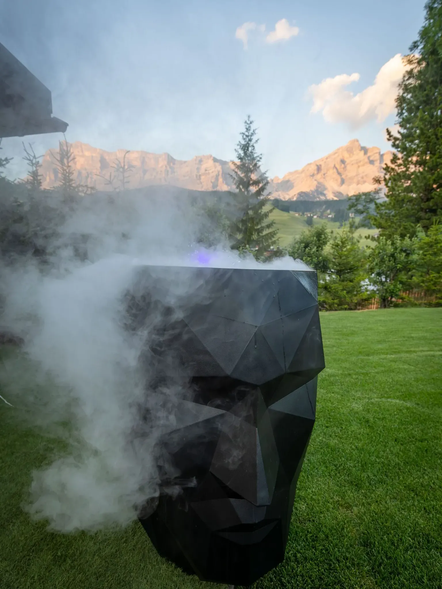 Smoky black geometric sculpture on green lawn with mountains and trees in background.