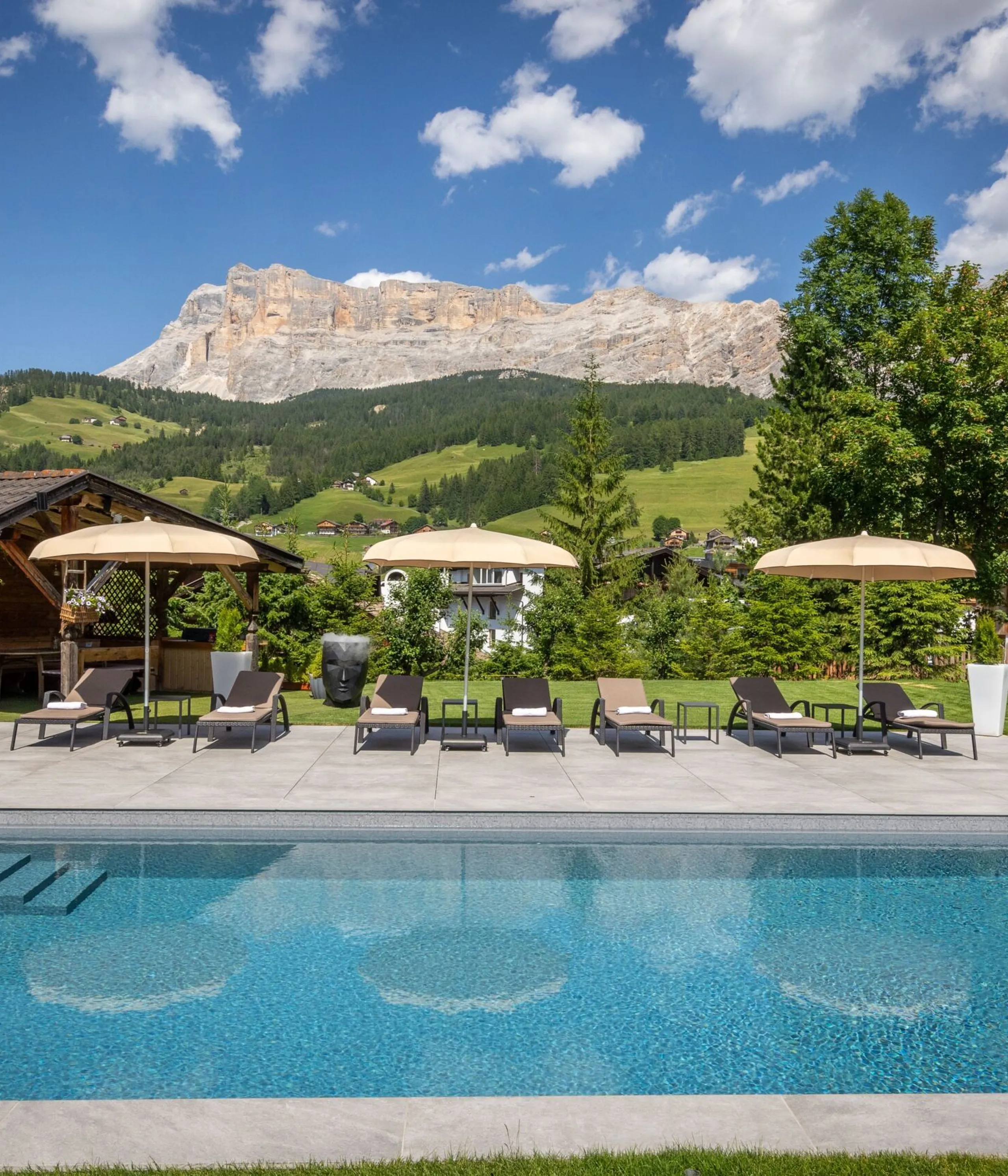 Outdoor pool area with lounge chairs and beige umbrellas, mountains and green hills in the background.