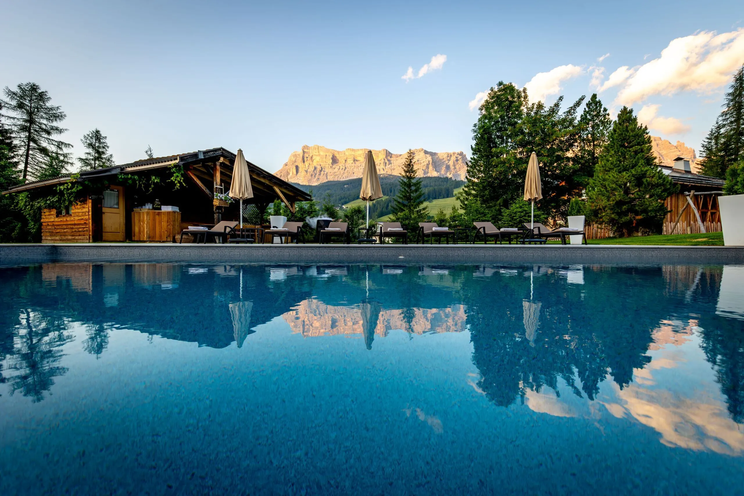 Sunlit outdoor pool area with wooden cabana, lounge chairs, and umbrellas, mountain backdrop reflecting in water.
