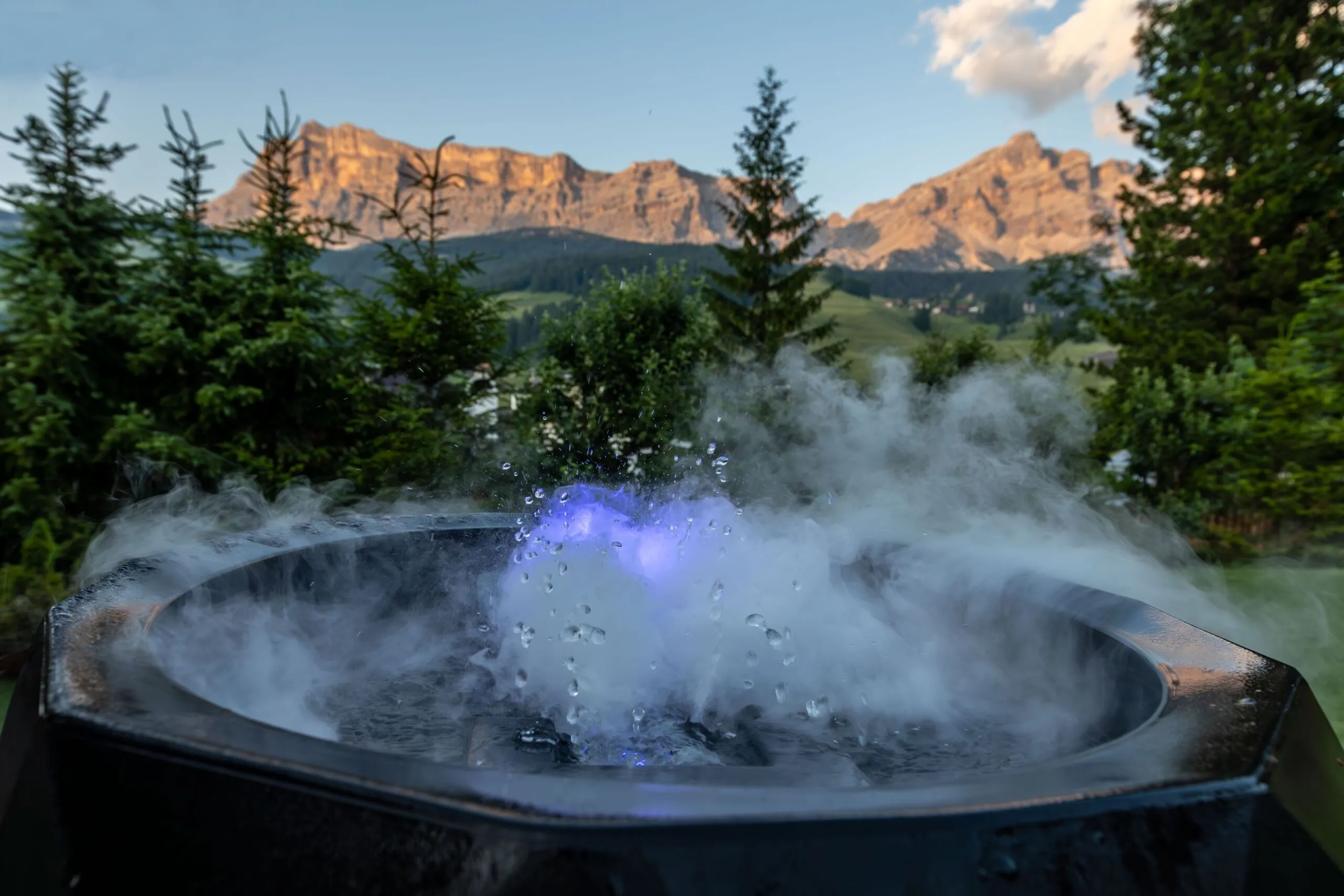 Steaming hot tub with rainbow glow, set against alpine trees and rugged mountains at sunset.