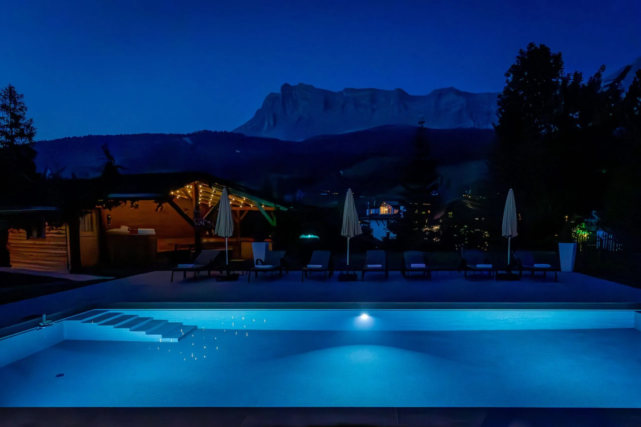Pool at night with blue lighting, lounge chairs and umbrellas along a patio, mountain range in the background.