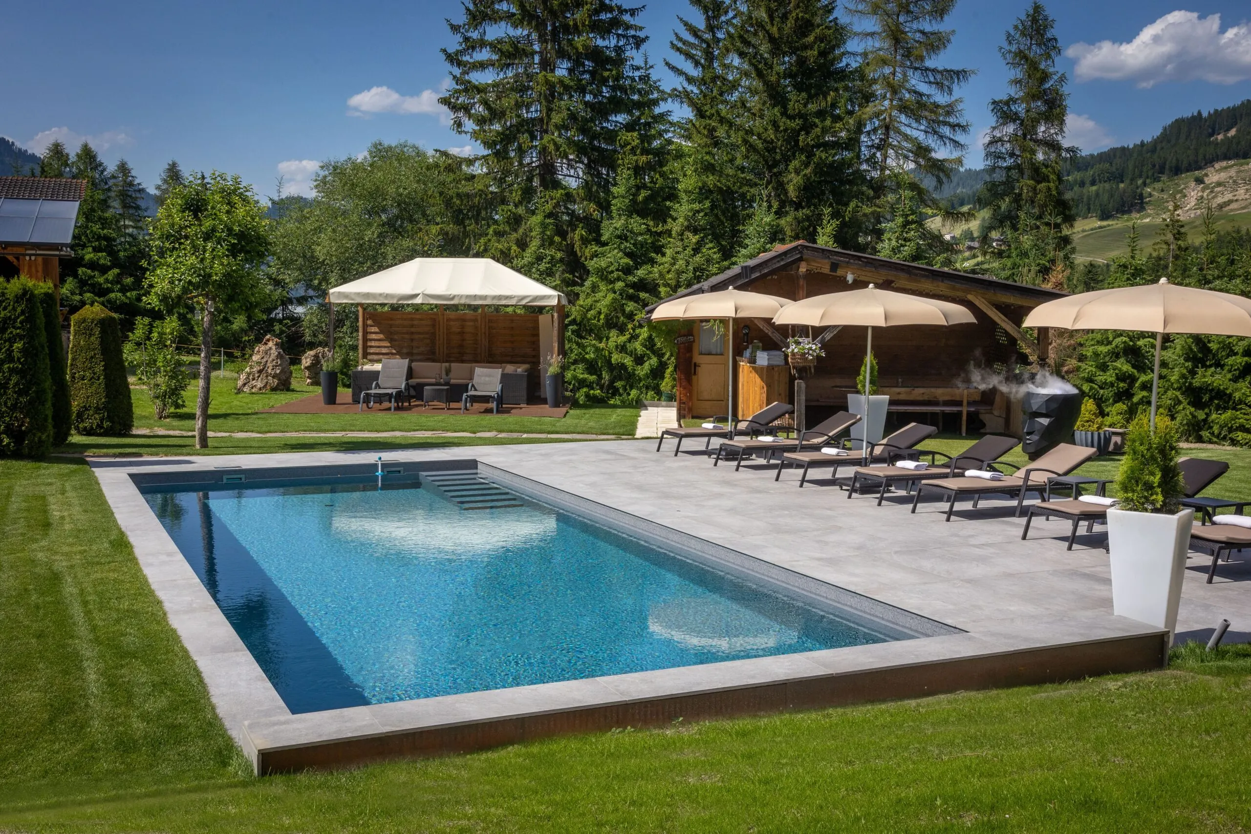 Luxurious outdoor pool area with lounge chairs, beige umbrellas, and wooden cabana set against green fields and pine trees.