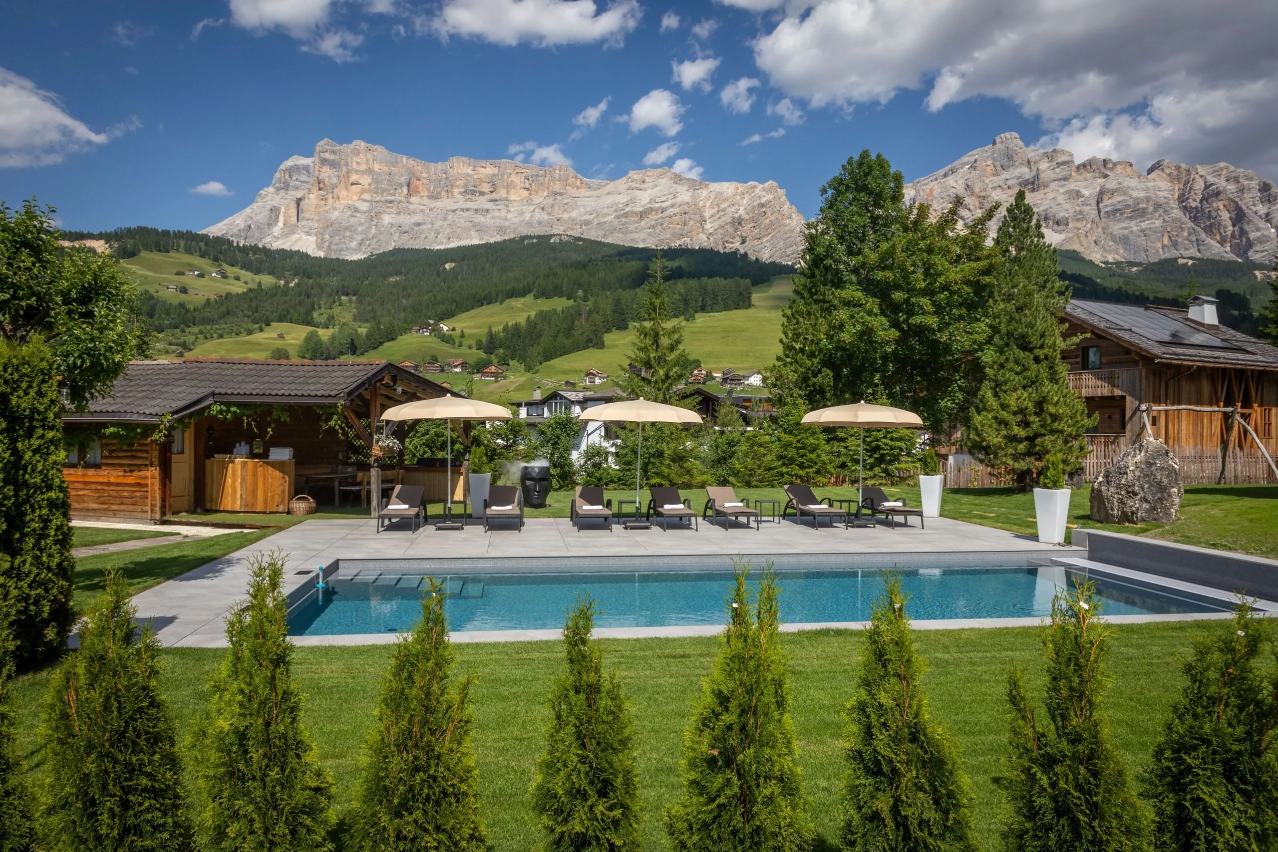 Outdoor luxury pool area with lounge chairs, umbrellas, wooden cabins, and alpine mountains in the background.