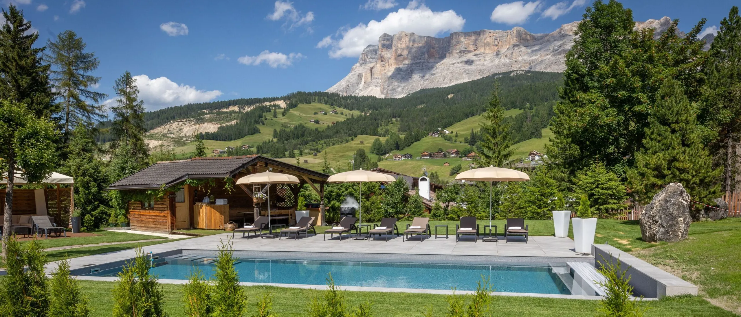 Outdoor pool area with lounge chairs, umbrellas, a wooden hut, and scenic green hills and mountains in the background.