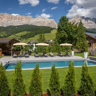 Outdoor pool area with loungers and umbrellas, wooden cabins, and Alpine mountains in the background.